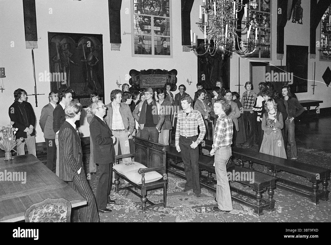 Ricevimento di studenti tedeschi Gravenzaal Reception, Whizgle News, Dutch Desk, Paesi Bassi, 1950 - 2000 il 16-03-1977. L'immagine contiene questi argomenti. La scena si sviluppa in una sala spaziosa e riccamente decorata caratterizzata dalla sua architettura storica. Le pareti sono rivestite da grandi finestre che consentono alla luce diffusa di diffondersi, illuminando delicatamente la stanza. Alte travi di legno si innalzano perpendicolarmente al soffitto, aggiungendo alla grandezza della camera. Sul pavimento, un tappeto di lusso dai toni profondi copre il terreno, contribuendo a creare un'atmosfera calda ma formale. Al centro, diversi individui si trovano in c Foto Stock