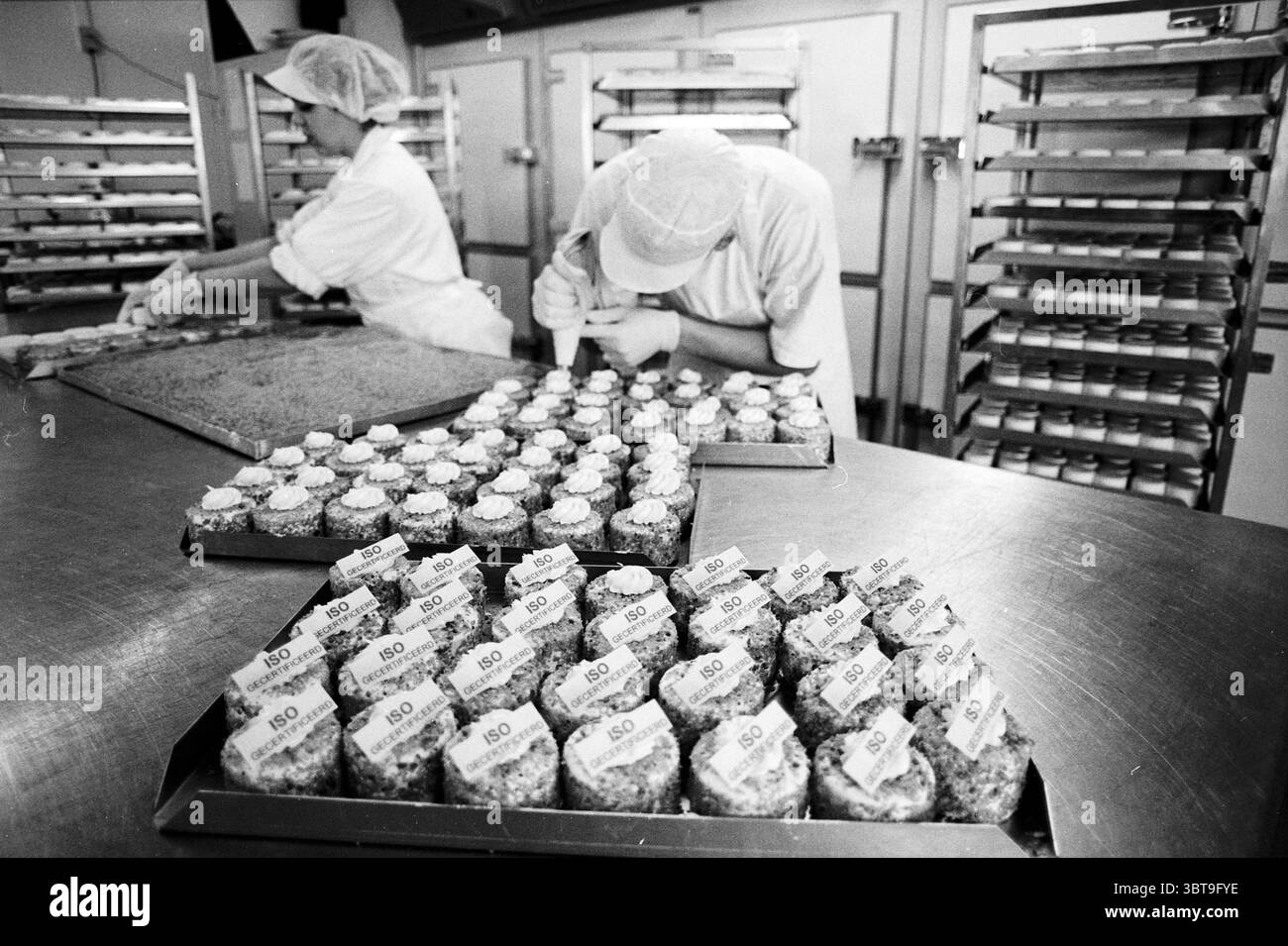 Pasticceria ISO standard PTT Post Amsterdam Amsterdam Paesi Bassi, Whizgle News, Dutch Desk, Paesi Bassi, 1950 - 2000 il 16-04-1997. Questi argomenti sono mostrati nell'immagine. In una cucina animata, due persone sono impegnate nel processo di cottura e decorazione dei dolci. La scena è dominata da un ambiente industriale, caratterizzato da superfici in acciaio inox e grandi vassoi da forno riempiti con una serie di dolcetti. In primo piano, spicca un vassoio di cupcake decorati in modo intricato. Ogni cupcake è decorato con un turbinio di cremosa glassa, creativamente condita per formare forme eleganti. Il Colo Foto Stock