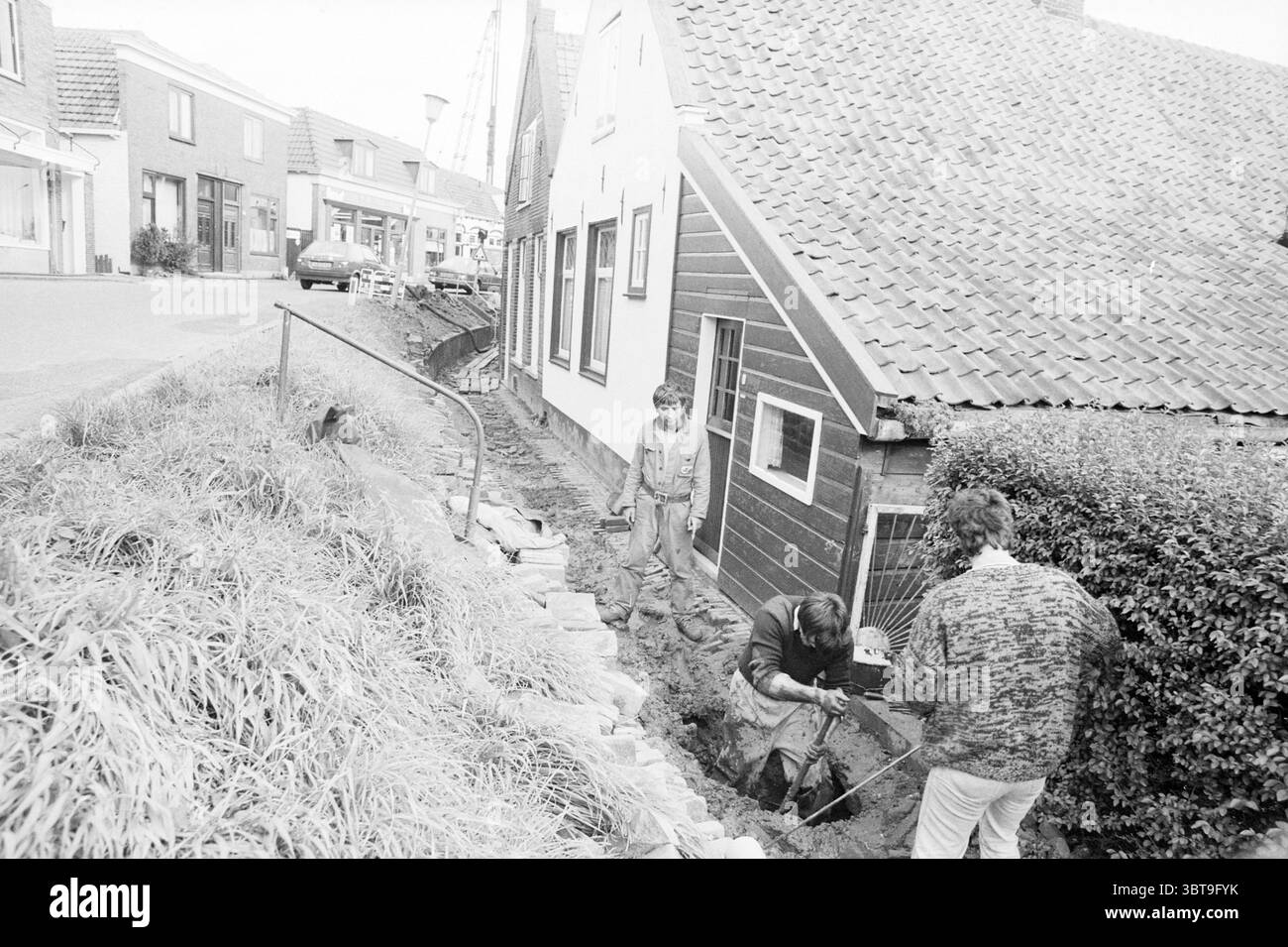 Impianti fognari Spaarndam Sewer Spaarndam, Whizgle News, Dutch Desk, Paesi Bassi, 1950 - 2000 il 09-10-1987. Questi sono gli argomenti nell'immagine. In una tranquilla zona residenziale, un paio di lavoratori è impegnato in un compito vicino alla fondazione di una pittoresca casa di legno. La casa presenta un distinto tetto inclinato ricoperto da un materiale testurizzato, e le sue pareti mostrano un'affascinante tonalità scura che contrasta con i toni più chiari degli edifici vicini. La scena è dominata da colori terrosi, con terreno esposto e ghiaia intorno ai lavoratori. Una persona è in una trincea poco profonda, concentrata intensamente su un'attività Foto Stock
