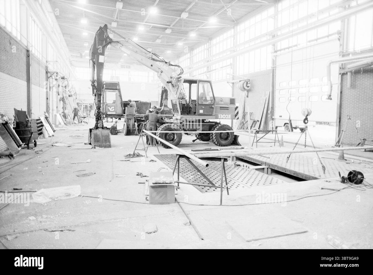 Costruzione del garage Vemat Cruquius. Vermeer Cruquius, Whizgle News, Dutch Desk, Paesi Bassi, 1950 - 2000 on 26-11-1987. Questi sono gli argomenti nell'immagine. La scena cattura una spaziosa officina industriale caratterizzata da una miscela di elementi in cemento e metallo. L'alto soffitto è rivestito da grandi e luminose luci che proiettano un'illuminazione forte nello spazio, creando un contrasto tra luce e ombra. In primo piano, un escavatore meccanico si trova in posizione prominente, con il braccio sollevato come se fosse pronto all'azione. Nelle vicinanze, un altro pezzo di macchinari pesanti è parcheggiato, suggerendo un flusso di lavoro Foto Stock