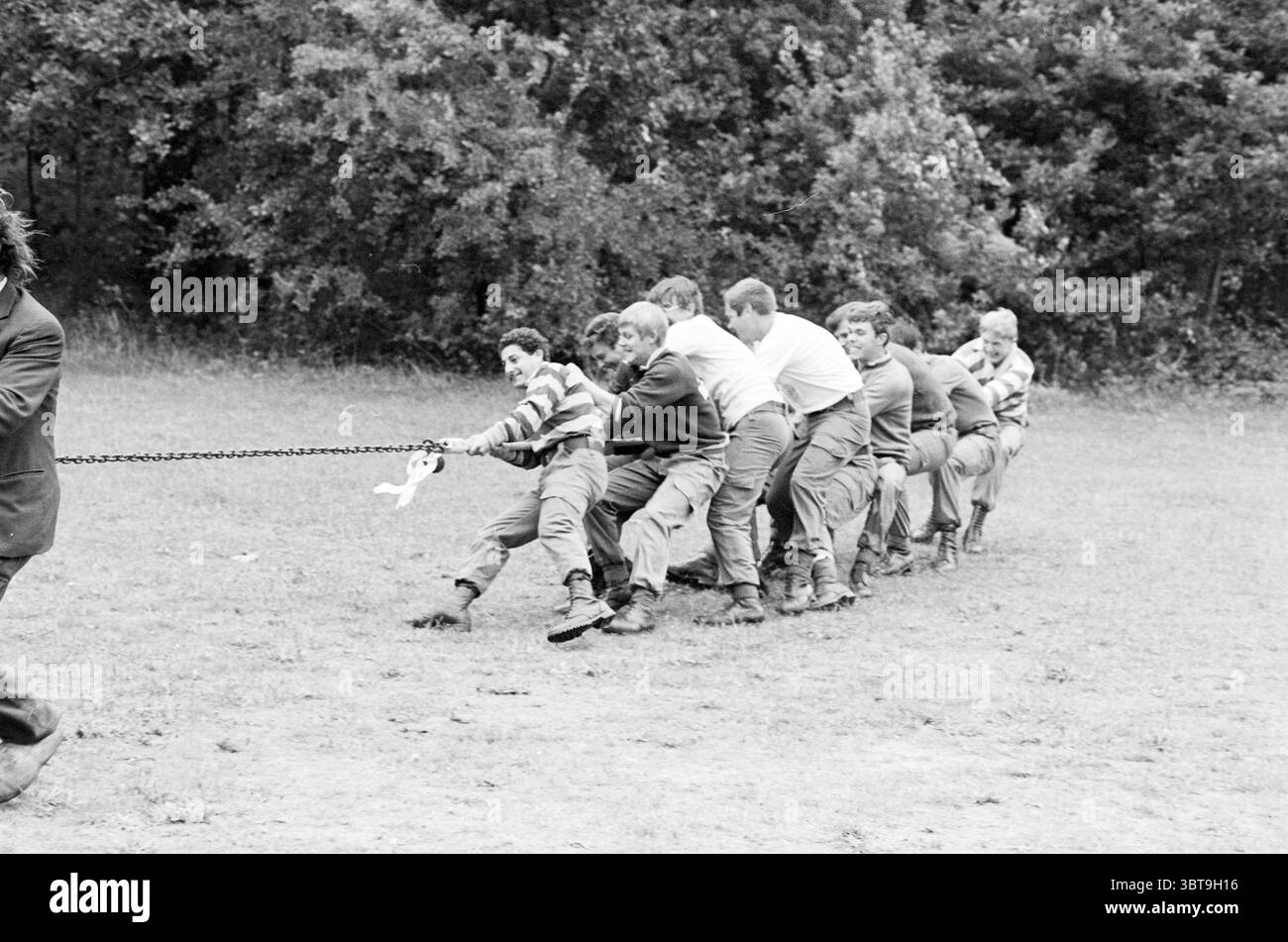 Rimorchiatore di guerra con cavallo dei fratelli SAS a IJmuiden Tug of War IJmuiden Paesi Bassi, Whizgle News, Dutch Desk, Paesi Bassi, 1950 - 2000 il 18-08-1982. L'immagine contiene questi argomenti. La scena raffigura un ambiente dinamico all'aperto in cui un gruppo di individui è impegnato in un'attività competitiva. In primo piano, viene tirata una corda chiara, indicando un tiro alla fune. I partecipanti sono allineati in fila, ciascuno tenendo la corda e appoggiandosi all'indietro con uno sforzo palpabile. Le loro espressioni mostrano un mix di concentrazione e determinazione, suggerendo l'intensità della competizione. La dentro Foto Stock