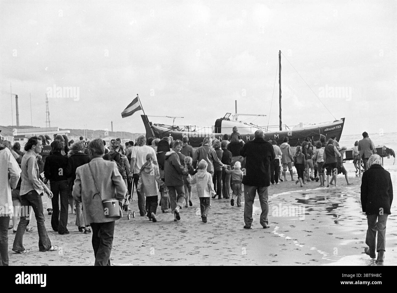 Esercizio KNRM Wijk aan Zee esercizio Wijk aan Zee, Whizgle News, Dutch Desk, Paesi Bassi, 1950 - 2000 il 25-07-1975. L'immagine contiene questi argomenti. La scena cattura un vivace ambiente di spiaggia, dove un grande gruppo di persone è radunato lungo la costa. Gli individui, vestiti con un mix di abbigliamento casual tipico dell'epoca, creano un arazzo di colori e stili, che spazia dai colori pastello chiari alle tonalità più scure. Alcuni portano l'attrezzatura da spiaggia, mentre altri si tengono per mano o si godono conversazioni informali. In primo piano, la spiaggia sabbiosa risplende di riflessi del cielo coperto, che è pieno di spirito Foto Stock