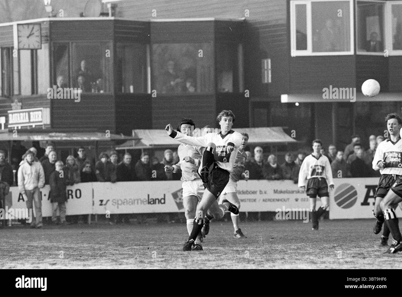 Calcio: HFC-Old Internationals Haarlem Paesi Bassi, Whizgle News, Dutch Desk, Paesi Bassi, 1950 - 2000 il 31-12-1992. L'immagine include questi argomenti. La scena cattura un momento vivace su un campo da calcio, dove un giocatore, vestito con un kit leggero, sta eseguendo un passaggio. Il giocatore mostra un'espressione determinata, i muscoli tesi, mentre manda abilmente la palla in volo. Sullo sfondo, una figura rivestita in nero si erge con una postura impaziente, pronta ad intercettare o a rispondere allo svolgimento del gioco. Il campo in sé è fangoso, con patch visibili che mostrano la i. Foto Stock