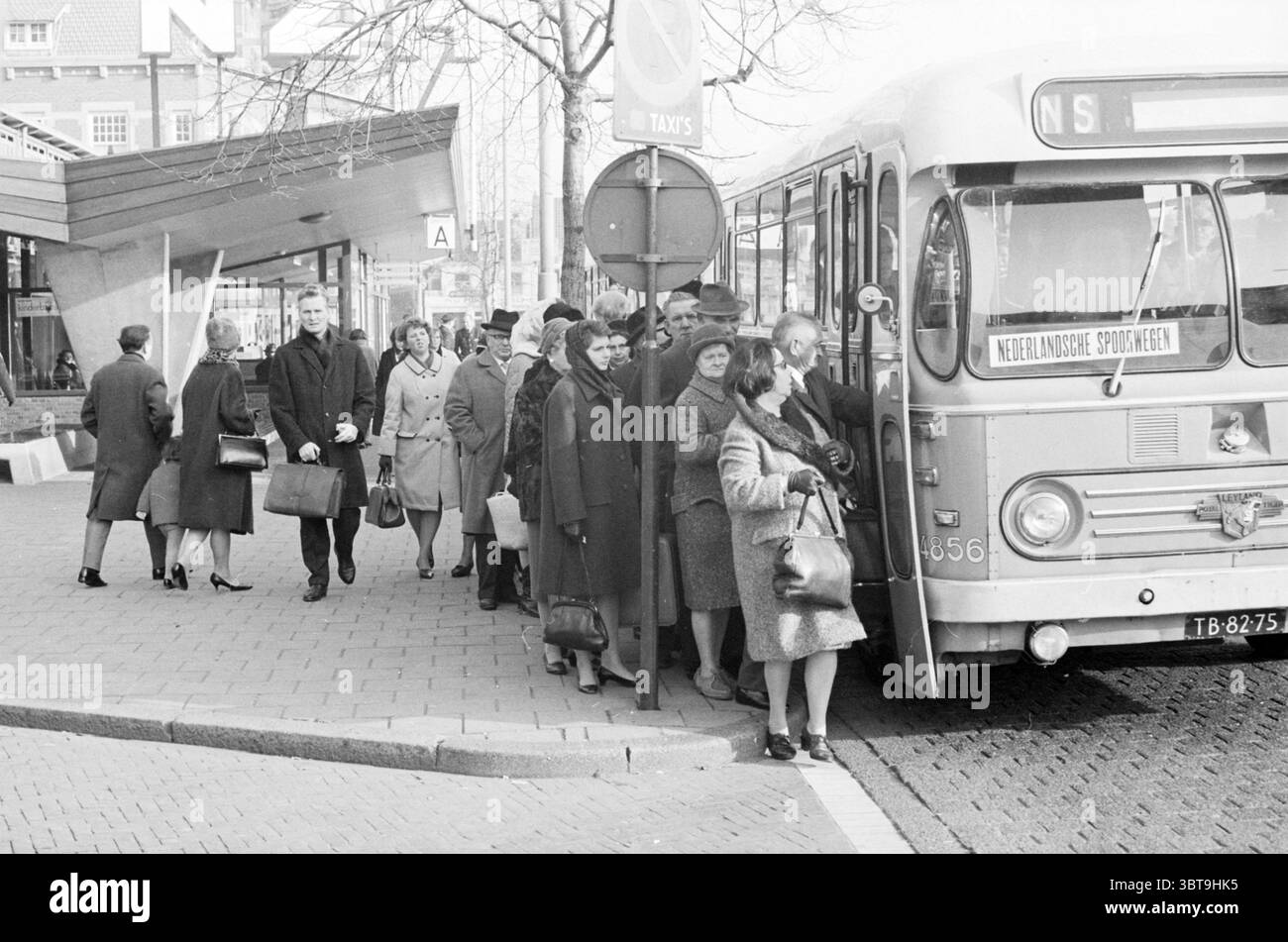 Autobus NZH utilizzato come trasporto temporaneo per i passeggeri del treno Haarlem Paesi Bassi., Whizgle News, Dutch Desk, Paesi Bassi, 1950 - 2000. Questi argomenti sono mostrati nell'immagine. Una frenetica scena urbana si svolge presso una fermata dell'autobus con un senso di movimento e attività. In primo piano, un gruppo di persone è riunito intorno a un vecchio autobus, che è parcheggiato con la porta aperta, invitando i passeggeri a salire a bordo. Le persone sono vestite con un mix di abbigliamento invernale formale e informale, prevalentemente in sfumature di grigio, nero e marrone. Molti indossano cappotti e cappelli, suggerendo una giornata fredda. Alcuni individui tengono in mano borse o SM Foto Stock