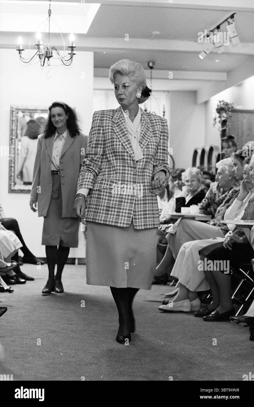Sfilata di moda, Whizgle News, Dutch Desk, Paesi Bassi, 1950 - 2000 nel 1995. Questi sono gli argomenti nell'immagine. La scena presenta una vivace sfilata di moda che si svolge al chiuso, caratterizzata da un ambiente luminoso e ben illuminato. La pista è centrale, fiancheggiata da spettatori seduti, molti dei quali sono donne anziane, indicando un pubblico maturo. Sono vestiti con una varietà di abiti eleganti e colorati, che suggeriscono un senso di occasione e stile. All'avanguardia, due modelli percorrono la pista. Il primo modello, che si muove con sicurezza in avanti, indossa un blazer sartoriale a quadri su una morbida blusa e una matita sk Foto Stock