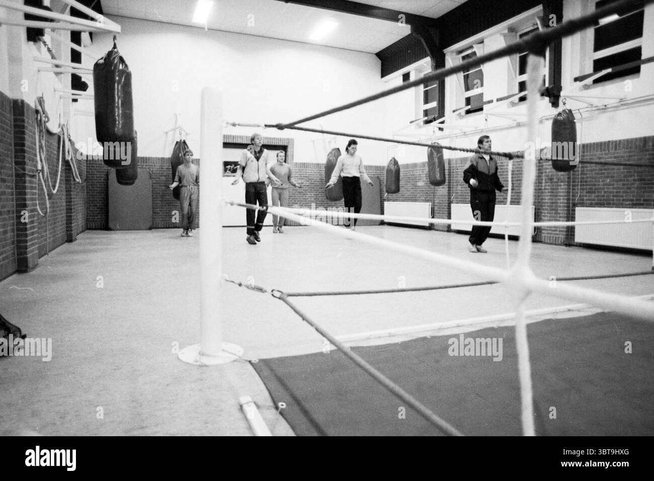 Boxing club de Ring Eksterstraat IJmuiden Boxing IJmuiden Nederland, Whizgle News, Dutch Desk, Paesi Bassi, 1950 - 2000 il 14-03-1989. L'immagine include questi argomenti. La scena cattura una palestra di boxe con un forte senso di movimento e attività. In primo piano, un anello di boxe bianco è contrassegnato da corde che si estendono lungo la scena, conducendo l'occhio verso due individui che camminano lungo il bordo dell'anello. Le loro posture sono rilassate e allo stesso tempo decise, suggerendo una conversazione informale o un cameratismo. Lo sfondo è caratterizzato da diversi sacchetti di punzonatura pesanti sospesi dal c Foto Stock
