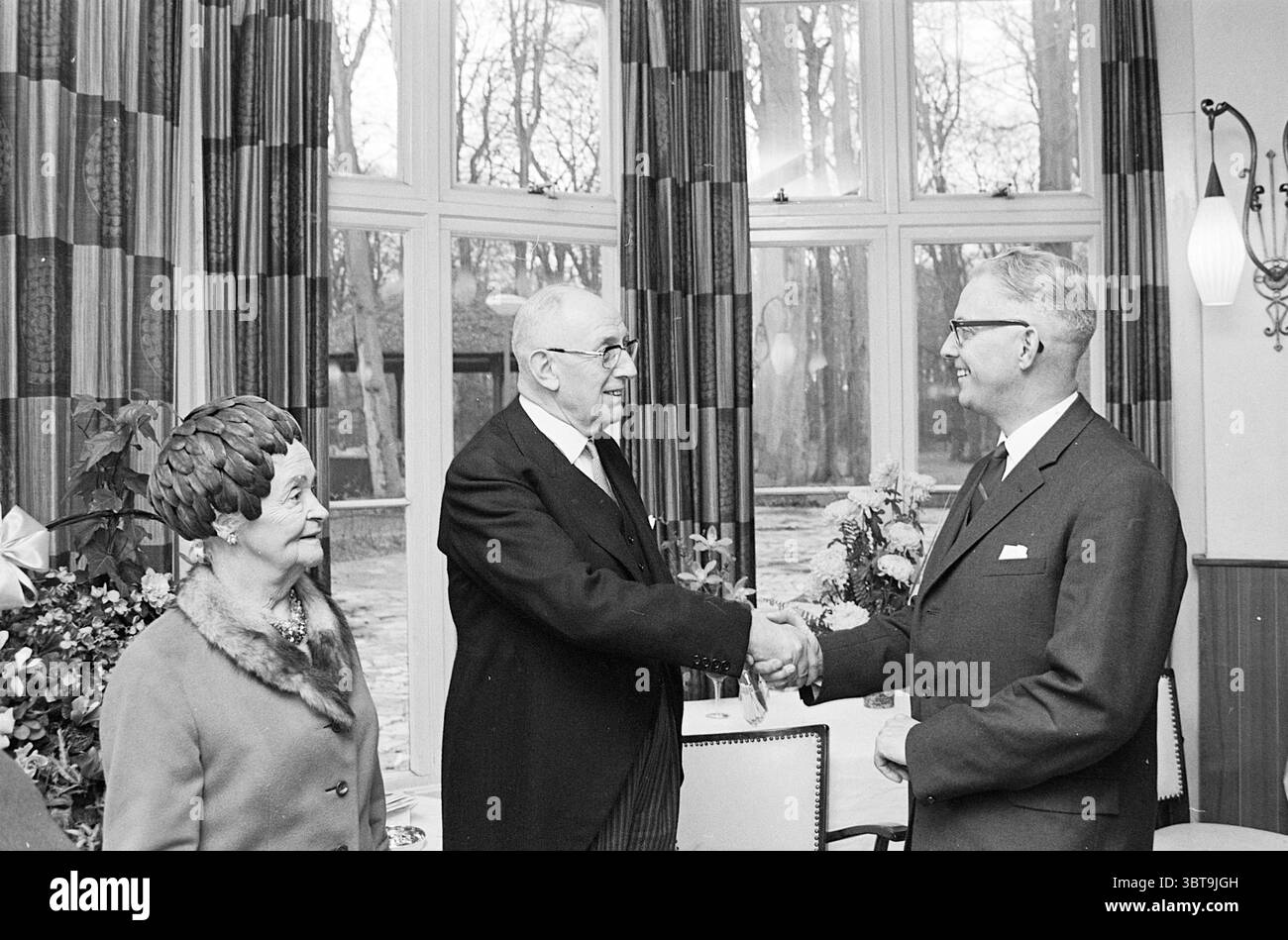 Addio Sig. Hortensius Noord Zuid Hollandse Vervoersmaatschappij N.V. NZ, Whizgle News, Dutch Desk, Paesi Bassi, 1950 - 2000 il 19-12-1967. L'immagine mostra questi argomenti. La scena cattura un'interazione formale tra tre individui in un ambiente interno che trasuda un'aria di importanza e dignità. In primo piano, due uomini sono impegnati in una stretta di mano, che significa un momento di accordo o di saluto. Un uomo, più anziano e dai capelli grigi, indossa un abito scuro con una camicia bianca e una cravatta, che proietta un senso di autorità e rispetto. La sua espressione è seria ma ottimista, suggerendo un Foto Stock