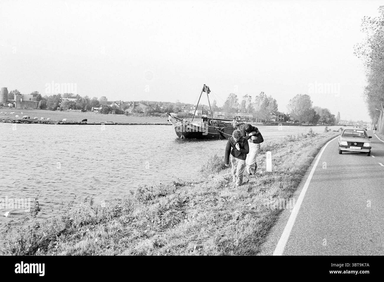 Tjalk attraverso Ringvaart presso Bennebroek strontrace Ships Competitions Bennebroek, Whizgle News, Dutch Desk, Paesi Bassi, 1950 - 2000 il 18-10-1983. Questi argomenti sono mostrati nell'immagine. La scena è caratterizzata da un tranquillo ambiente lungo il fiume, dove l'acqua splende dolcemente sotto la luce soffusa del giorno. Una grande barca, forse una chiatta, è ancorata nelle vicinanze, con una bandiera che oscilla leggermente nella brezza. La costa è costeggiata da lussureggiante erba verde che si fonde perfettamente con l'acqua. In primo piano, due figure sono impegnate in qualche attività, magari esplorando o raccogliendo qualcosa da terra. Il loro casual Foto Stock