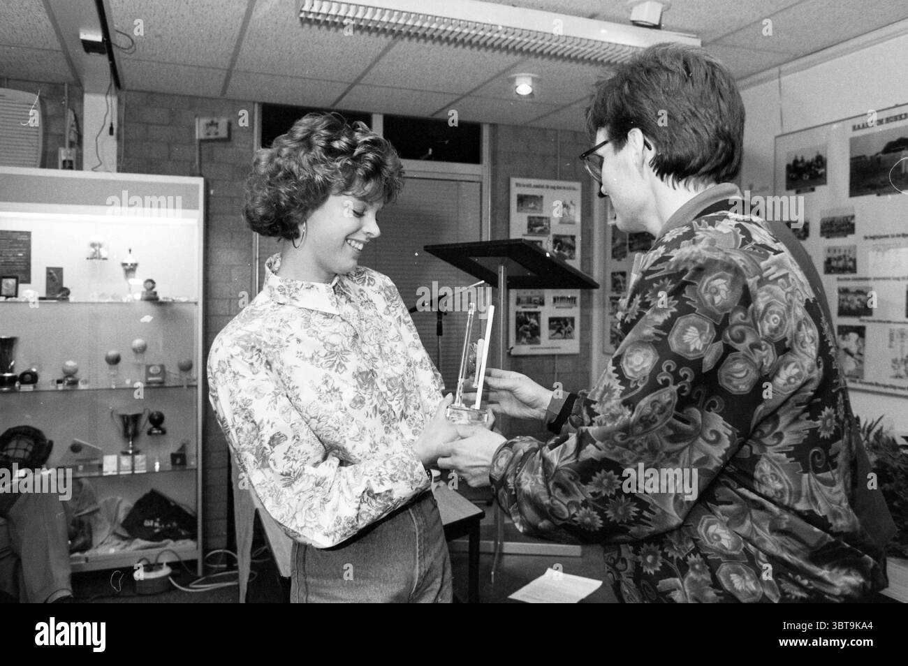 Cerimonia di premiazione del baseball e del softball 1990, Whizgle News, Dutch Desk, Paesi Bassi, 1950 - 2000 il 08-03-1991. Questi argomenti sono mostrati nell'immagine. In un ambiente interno ben illuminato, due persone sono impegnate in un momento di festa e riconoscimento. Una persona, una giovane donna con i capelli ricci, appare gioiosa quando riceve un premio. Indossa una blusa a motivi floreali che trasmette un senso di vivacità. I colori del suo vestito sono morbidi e pastello, contribuendo a creare un'atmosfera allegra. L'altra persona, presumibilmente un'autorità o un mentore, sta consegnando un trofeo, Foto Stock