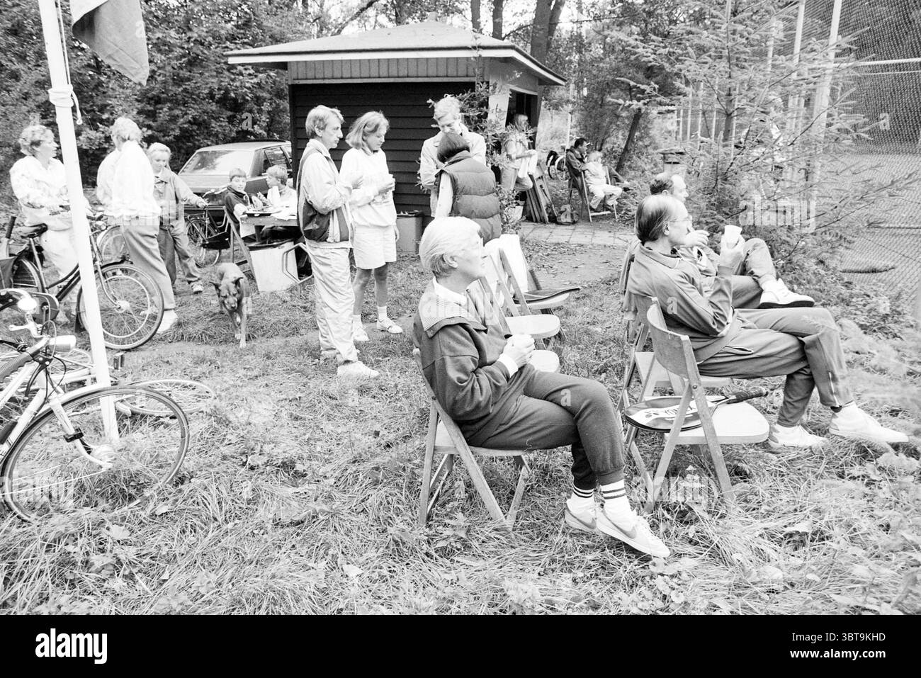 Alcune persone nei campi del tennis club, Whizgle News, Dutch Desk, Paesi Bassi, 1950 - 2000 il 07-10-1989. Questi sono gli argomenti nell'immagine. La scena cattura un incontro informale all'aperto, adagiato su uno sfondo di verde e sfumature di terra. È possibile vedere un gruppo di persone, alcune sedute su sedie di legno disposte in modo rilassato, mentre altre si trovano nelle vicinanze, interagendo con l'ambiente circostante o conversando. I partecipanti sono vestiti con un abbigliamento estivo casual, che spazia da pantaloncini di colore chiaro a abbigliamento sportivo, mostrando una varietà di motivi, alcuni sottili, mentre altri sono più vasti Foto Stock
