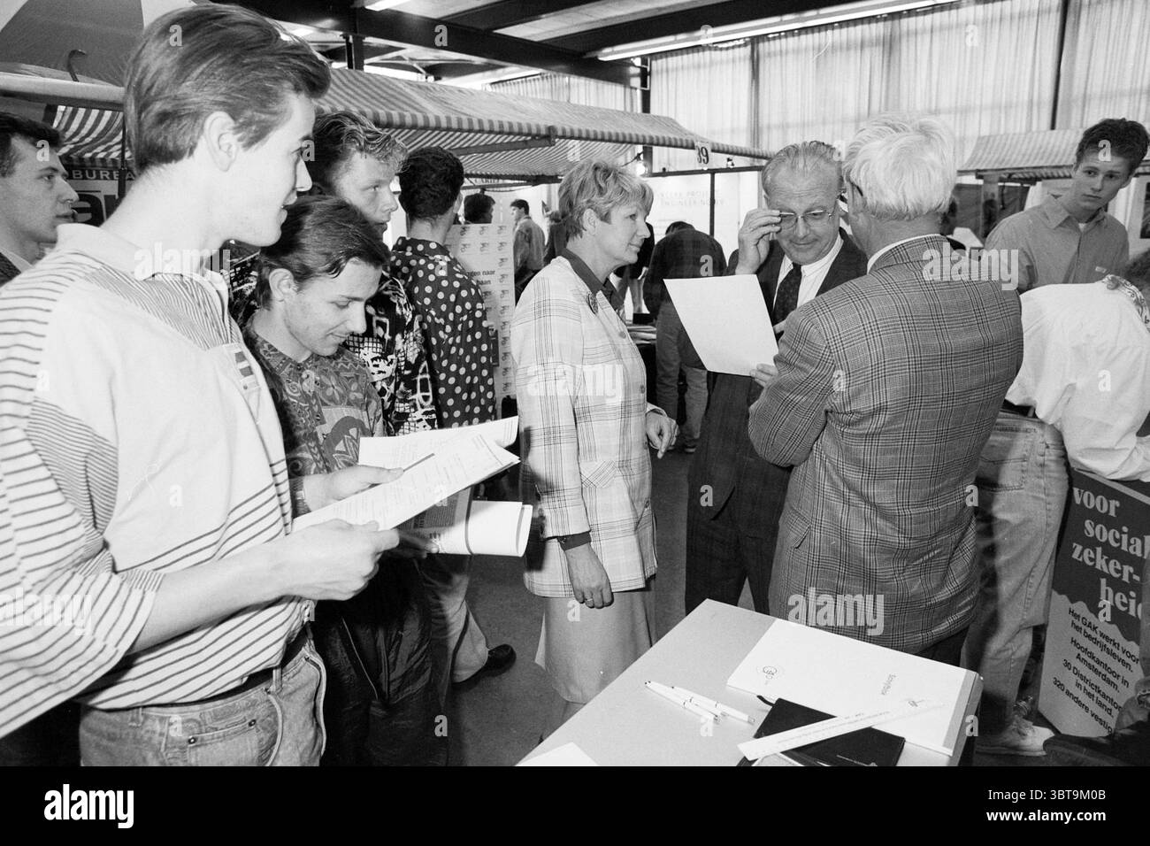 Il sindaco di IJmuiden apre il mercato del lavoro, Whizgle News, Dutch Desk, Paesi Bassi, 1950 - 2000 il 09-04-1992. L'immagine contiene questi argomenti. La scena è ambientata in un ambiente interno spazioso e ben illuminato, probabilmente una fiera o una sala esposizioni. L'illuminazione è luminosa, emettendo un bagliore naturale che esalta i colori vibranti degli abiti indossati dagli individui presenti. Un gruppo di persone è impegnato in una conversazione vicino a un tavolo ingombrante pieno di documenti e documenti. Ci sono cinque individui in primo piano, due dei quali sono vestiti con un abbigliamento leggermente formale, mentre altri indossano un cl più casual Foto Stock