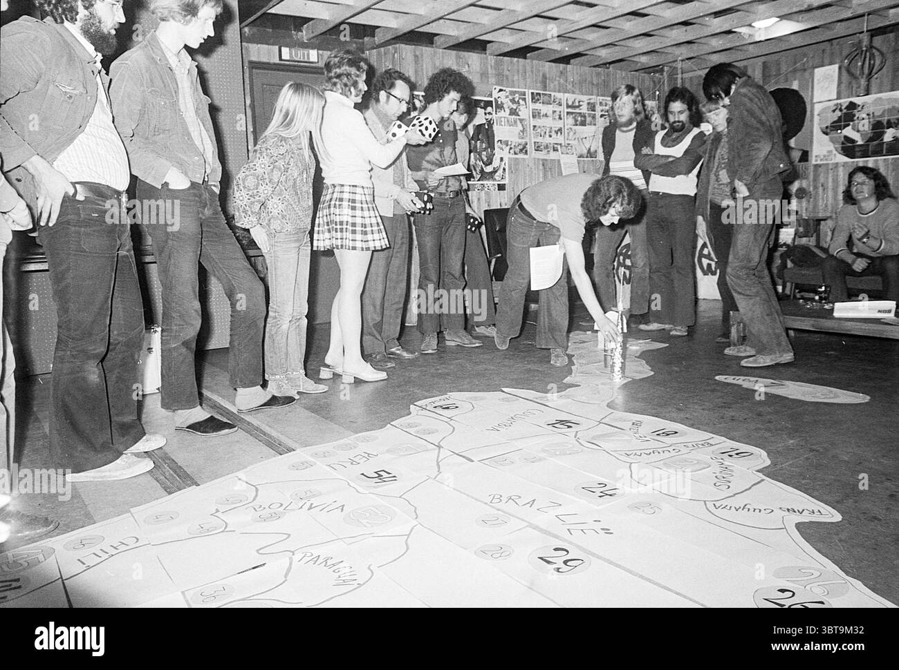 South American Goose giochi da tavolo e giocattoli, Whizgle News, Dutch Desk, Paesi Bassi, 1950 - 2000 il 26-09-1972. Questi sono gli argomenti nell'immagine. La scena è ambientata in una sala rustica e poco illuminata, probabilmente un centro comunitario o uno spazio di laboratorio, caratterizzata da travi in legno e da un semplice soffitto non adornato. Diversi gruppi di persone sono riuniti, impegnati in un'attività che sembra coinvolgere una grande mappa distribuita su tutto il pavimento, con varie aree delineate e numerate. In primo piano, una donna si inginocchia, concentrandosi intensamente sulla mappa mentre versa una sostanza da un contenitore. Il suo stile è CA Foto Stock