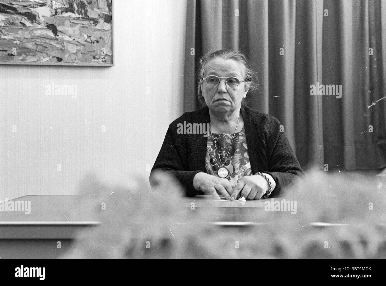 Sig.ra De Haan ex governante Menten Persons, Whizgle News, Dutch Desk, Paesi Bassi, 1950 - 2000 il 26-11-1976. L'immagine include questi argomenti. La scena raffigura una donna anziana seduta su un tavolo semplice, che trasmette un senso di quiete e introspezione. Ha un'espressione solenne, con occhi profondi che riflettono una vita di esperienze. I suoi capelli sono grigi e legati, rivelando il suo volto intemprato, che mostra segni di età, tra cui rughe e pieghe intorno alla bocca e alla fronte. Indossa un cardigan scuro su una blusa dai motivi vivaci che aggiunge un tocco di colore alla mut Foto Stock