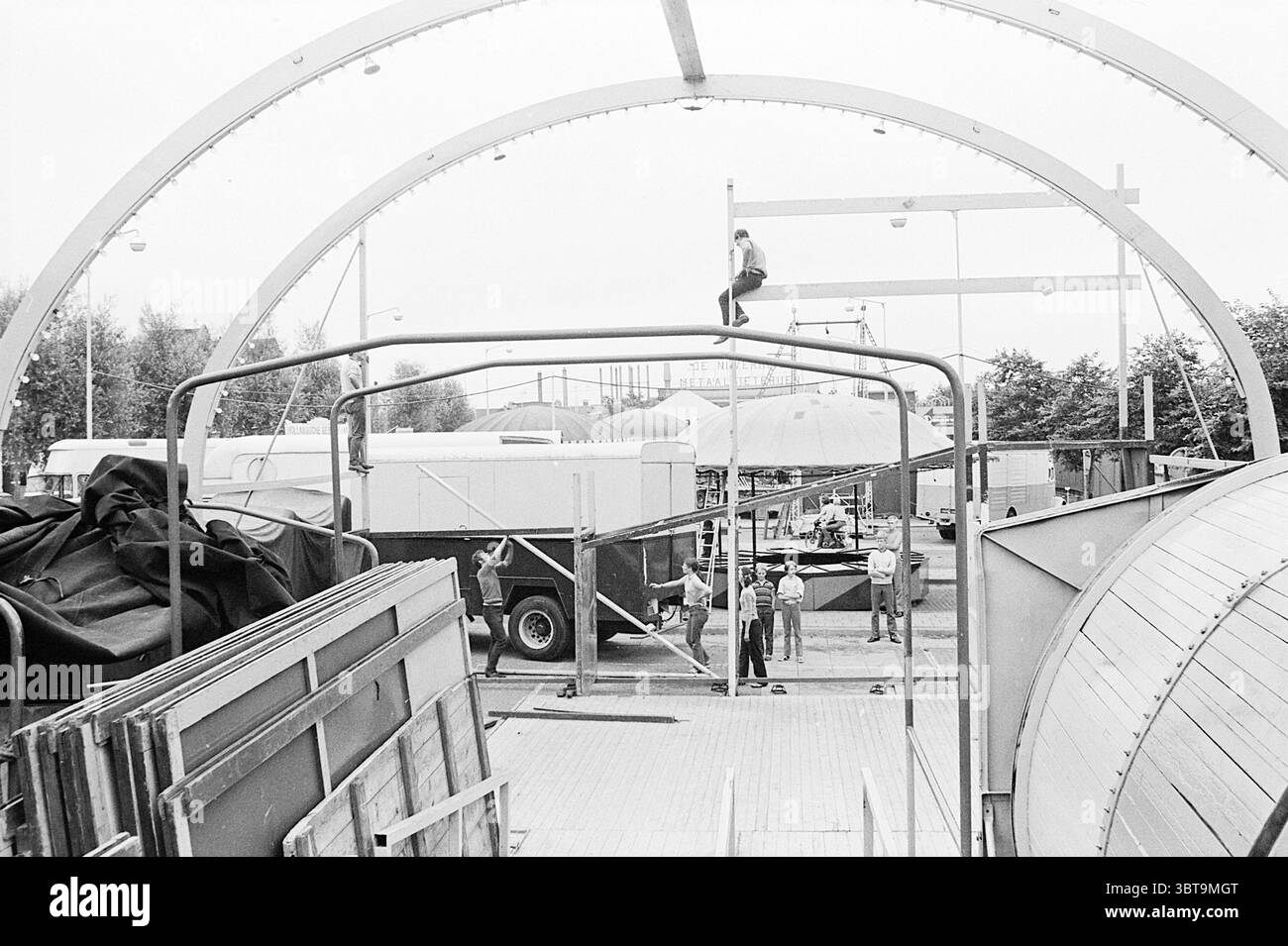 Festa per bambini Bakkum giochi vacanze vacanze vacanze vacanze vacanze campo, Whizgle News, Dutch Desk, Paesi Bassi, 1950 - 2000 il 31-07-1969. Questi sono gli elementi dell'immagine. La scena si sviluppa in una vivace area di costruzione incorniciata da archi che creano una struttura semi-aperta. I colori dominanti sono silenziati, con un tono monocromatico che aggiunge una sensazione nostalgica, quasi industriale. In primo piano, pile di pannelli metallici e teloni pesanti sono disposti in modo casuale, suggerendo il lavoro in corso e un ambiente caotico ma deciso. Le loro diverse texture aggiungono interesse, contrastando il metallo liscio del Foto Stock
