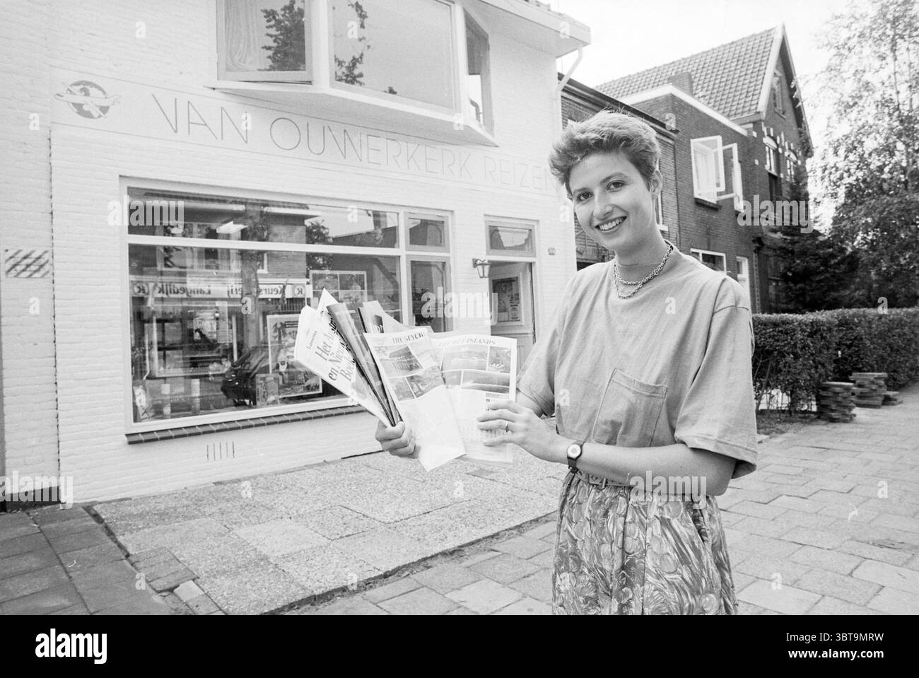 Van Ouwerkerk Reizen Santpoort Reizen Santpoort, Whizgle News, Dutch Desk, Paesi Bassi, 1950 - 2000 il 15-08-1989. L'immagine contiene questi argomenti. In un ambiente urbano vivace, una giovane donna si trova in primo piano con fiducia, tenendo in mano un pacchetto di opuscoli di viaggio. È vestita con una camicia leggermente oversize di colore chiaro e una gonna a motivi geometrici, che aggiunge un tocco di vivacità alla scena. I suoi capelli sono modellati con un taglio corto e alla moda. Lo sfondo rivela un edificio bianco ben tenuto con grandi finestre, suggerendo un'agenzia di viaggi, come dimostrano le brochure che ha in mano. L'ambiente circostante Foto Stock