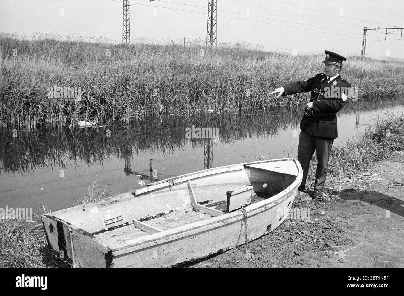 Agente di polizia nel fosso di Hillegom Police Hillegom, Whizgle News, Dutch Desk, Paesi Bassi, 1950 - 2000 il 04-06-1973. Questi argomenti vengono visualizzati nell'immagine. Una figura si trova vicino al bordo di un calmo corpo d'acqua, gestendo con una mano qualcosa di invisibile. Vestito con un'uniforme scura con un berretto, la persona sembra essere un poliziotto o una figura autoritaria. Il tessuto morbido dell'uniforme contrasta con la rugosità dell'ambiente naturale. A sinistra, una vecchia barca di legno è parzialmente spiaggiata sulla riva fangosa, la sua vernice scheggiata e sbiadita. La struttura della barca mostra segni di usura, con ope Foto Stock