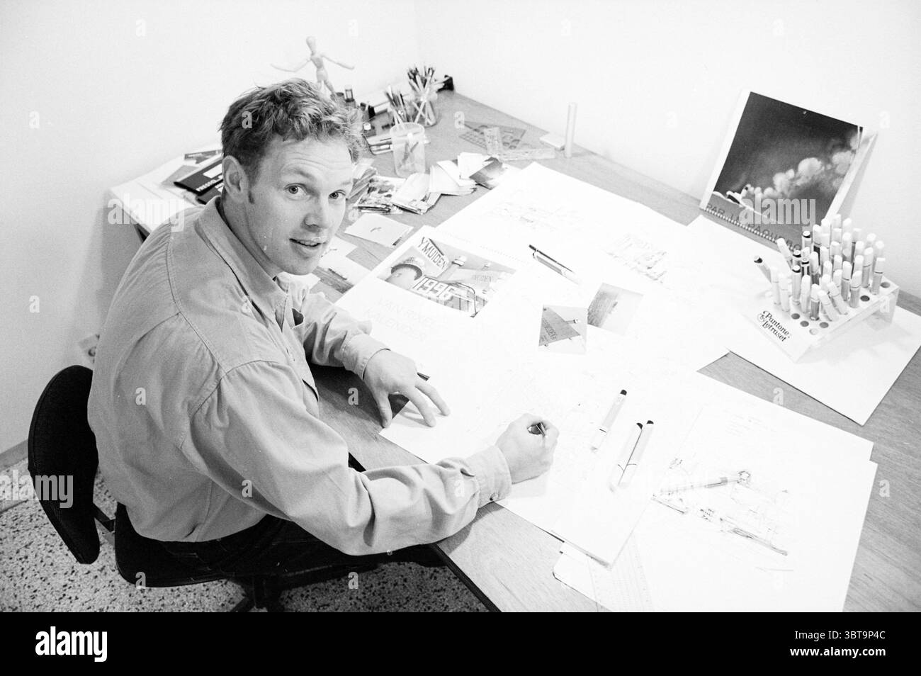 Artista Marco Stoker H'lem Haarlem Paesi Bassi, Whizgle News, Dutch Desk, Paesi Bassi, 1950 - 2000 il 26-10-1995. Questi argomenti sono mostrati nell'immagine. La scena cattura uno spazio di lavoro pieno di energia creativa. Un uomo siede su una scrivania di legno, impegnato nel disegno o nello schizzo, con vari strumenti sparsi intorno a lui. Indossa una camicia di colore chiaro, e la sua espressione è concentrata ma accessibile, suggerendo una miscela di concentrazione ed entusiasmo. Sparsi per la scrivania ci sono fogli di carta riempiti di intricati schizzi e disegni, che suggeriscono un impegno artistico. Diverse penne e pennarelli Foto Stock