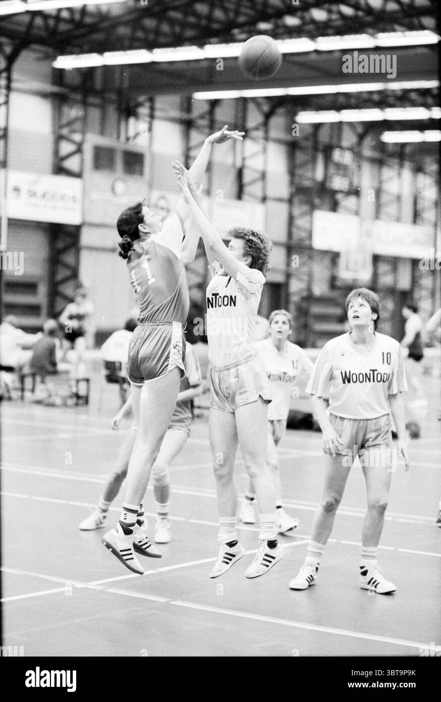 Partita di basket femminile., Whizgle News, Dutch Desk, Paesi Bassi, 1950 - 2000. L'immagine contiene questi argomenti. In una vivace palestra coperta, i giocatori sono attivamente impegnati in una partita di basket. La scena è dinamica e mostra un chiaro momento di competizione. In primo piano, due giocatori sono nel bel mezzo di un colpo di salto. Un atleta, vestito con un'uniforme grigia con un numero sul retro, salta per sparare la palla. L'altro, in uniforme bianca, sta raggiungendo per bloccare il colpo, con le braccia estese in alto sopra la testa. Entrambi i giocatori mostrano una concentrazione intensa, le loro espressioni trasmettono determinatio Foto Stock
