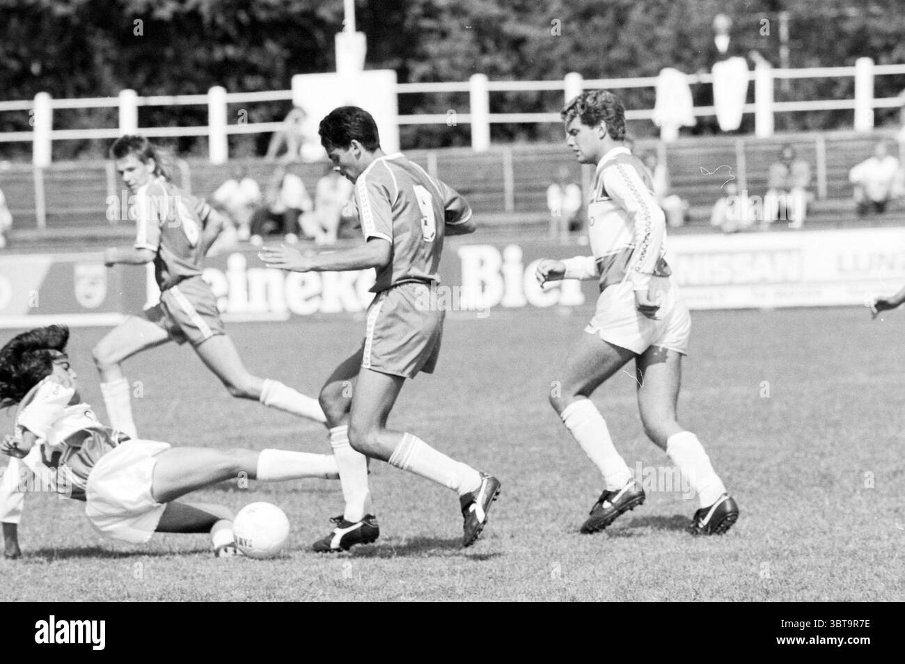 Torneo di calcio Ripperda Football, Whizgle News, Dutch Desk, Paesi Bassi, 1950 - 2000 il 13-08-1989. Questi sono gli elementi dell'immagine. La scena cattura un momento di energia da una partita di calcio su un ampio campo erboso. In primo piano, due giocatori sono impegnati in un gioco intenso. Un giocatore, vestito con un'audace maglia rossa, sta manovrando abilmente la palla mentre si trova di fronte a un avversario con un'uniforme bianca. La divisa bianca presenta sottili strisce che enfatizzano il contrasto tra le due squadre. L'erba è un verde vibrante, a testimonianza della buona tenuta del surf Foto Stock