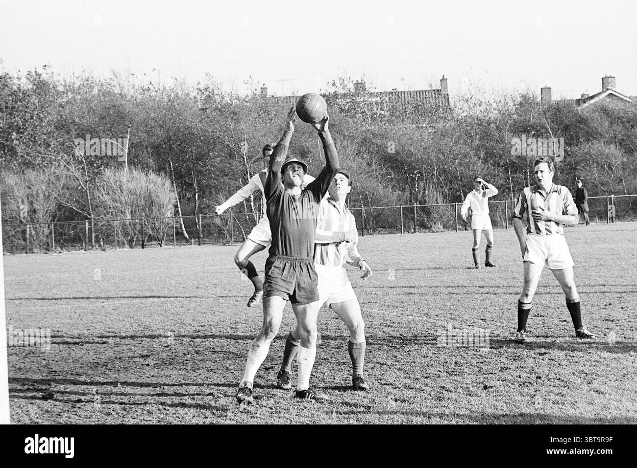 Waterloo - IEV Football, Whizgle News, Dutch Desk, Paesi Bassi, 1950 - 2000 il 07-11-1965. Questi sono gli argomenti nell'immagine. La scena cattura un momento intenso durante una partita di calcio su un campo erboso, con i giocatori impegnati attivamente nel gioco. Un portiere, che indossa una maglia scura e pantaloncini corti, si allunga verso l'alto per catturare un pallone, mostrando determinazione e atleticità. La sua postura suggerisce che è a metà passo, con un braccio completamente esteso e le dita che si avvicinano alla palla, che si trova appena sopra la sua testa. Intorno a lui, anche molti altri giocatori sono in movimento. Due avversari sono molto vicini alla posizione Foto Stock