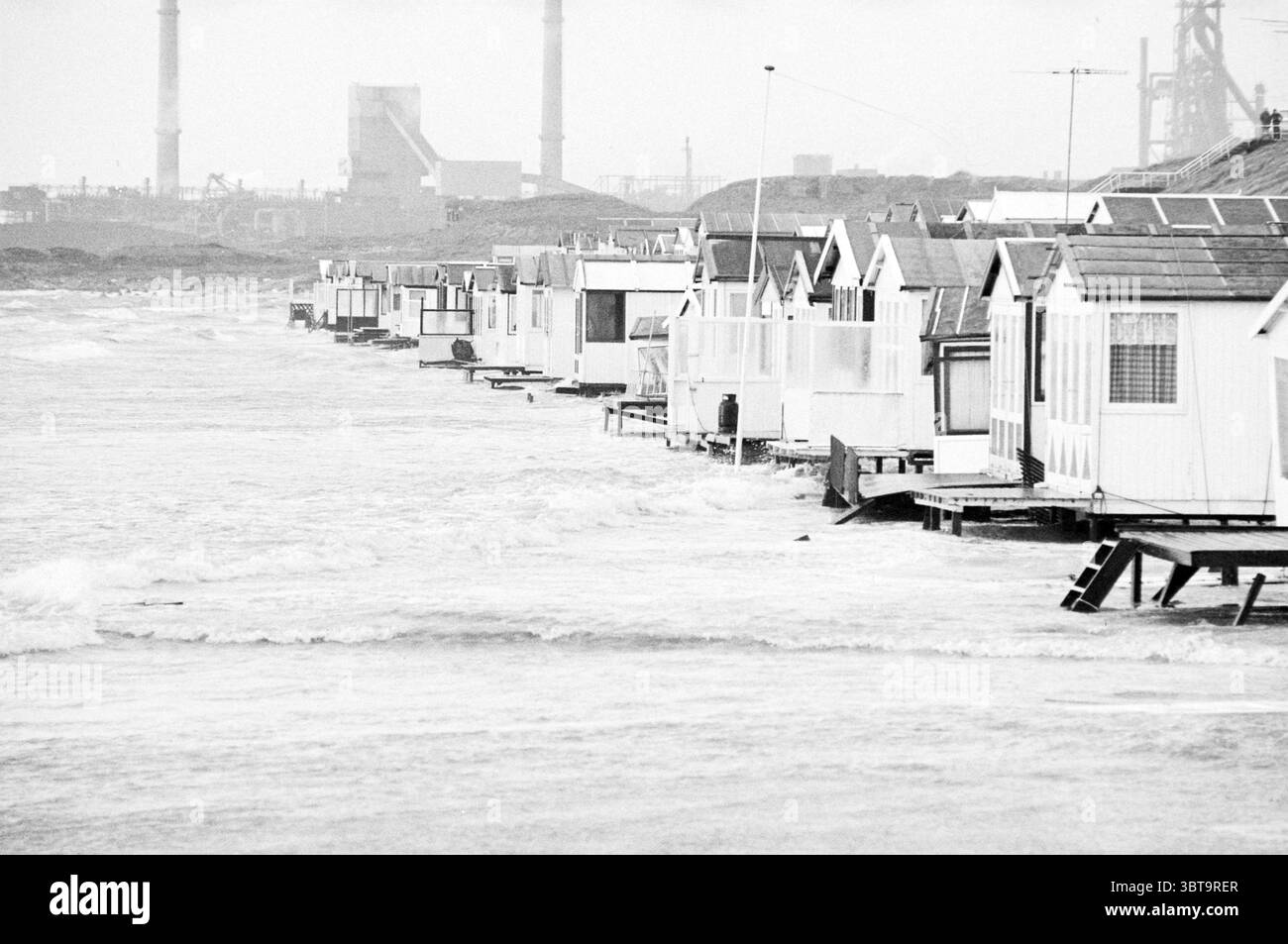 Tempesta spiaggia IJmuiden tempesta e danni tempesta Spiaggia e spiagge IJmuiden Paesi Bassi, Whizgle News, Dutch Desk, Paesi Bassi, 1950 - 2000 il 19-04-1980. Questi argomenti sono mostrati nell'immagine. La scena cattura un ambiente costiero dove una fila di piccole capanne sulla spiaggia si allinea lungo la costa. Queste capanne, dipinte in bianco tenue e colori pastello chiari, hanno tetti inclinati e grandi finestre, che donano loro un aspetto pittoresco e affascinante. Il tempo è coperto, emettendo un tono di grigio attenuato sull'ambiente, con nuvole scure che incombono in alto. Le onde si infrangono delicatamente ai bordi delle capanne, suggerire Foto Stock