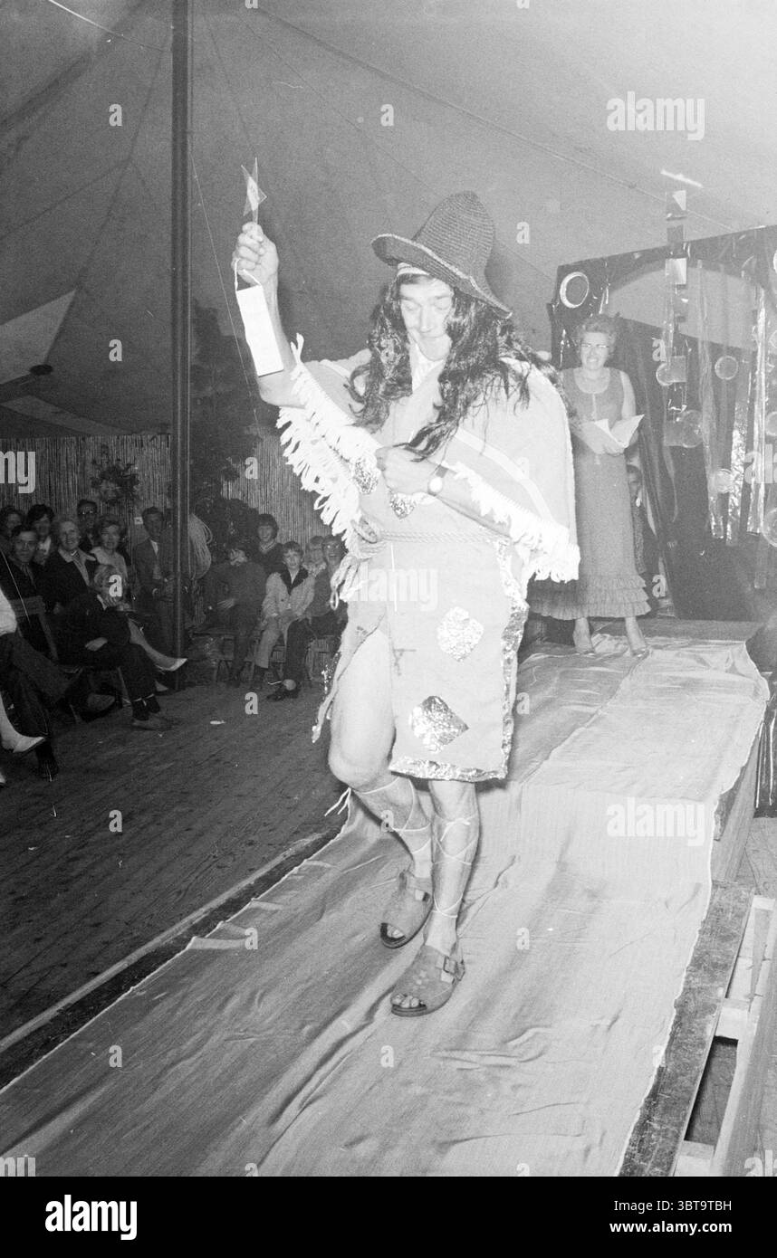 Sfilata di moda Rijsenhout sfilata di moda, Whizgle News, Dutch Desk, Paesi Bassi, 1950 - 2000 il 07-06-1971. Questi sono gli argomenti nell'immagine. La scena si sviluppa all'interno di una spaziosa tenda, con i suoi tessuti drappeggiati e decorati, creando un'atmosfera festosa. Una piattaforma simile a una pista si estende dal primo piano verso lo sfondo, dove il pubblico siede, con un coinvolgimento attento. La folla, composta da uomini e donne, è vestita in una varietà di stili che vanno dal casual al semi-formale, mostrando un senso di diversità. Al centro della composizione, un esecutore percorre la pista, trasudando confido Foto Stock