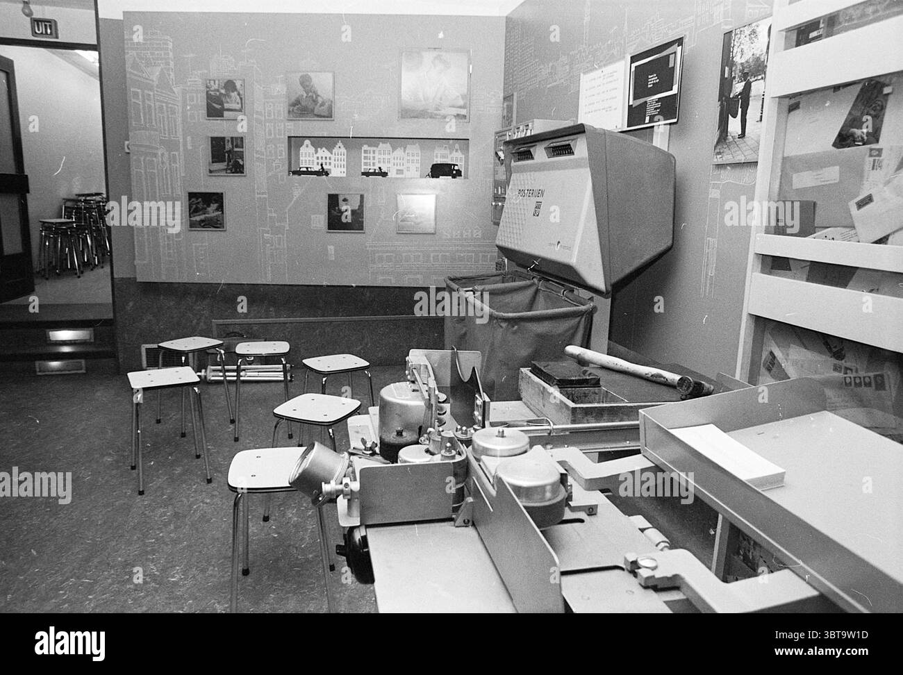 Mostra della posta P.T.T. PTT servizi postali City Post, Whizgle News, Dutch Desk, Paesi Bassi, 1950 - 2000 il 11-03-1974. Questi argomenti vengono visualizzati nell'immagine. La scena raffigura una stanza piena di elementi che evocano un senso di nostalgia e creatività. In primo piano, una macchina multifunzione domina lo spazio, le sue superfici metalliche riflettono una luce soffusa. Questa macchina ha una leggera lucentezza, indicativa di un uso regolare, con varie manopole e leve che ne indicano il funzionamento complesso. Intorno alla macchina sono presenti alcuni piccoli sgabelli minimalisti dal design semplice e funzionale Foto Stock