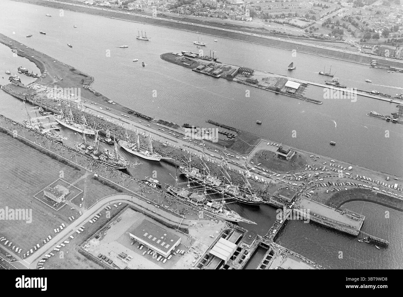 Fotografie aeree della partenza delle navi di Sail Amsterdam 1980 alle chiuse di IJmuiden. Per navigare. IJmuiden Paesi Bassi, Whizgle News, Dutch Desk, Paesi Bassi, 1950 - 2000 il 12-08-1980. L'immagine include questi argomenti. La scena presenta un vivace porto pieno di attività. Al centro, numerose navi sono attraccate insieme, i loro alberi raggiungono l'alto come una foresta di legno e vele. Le navi variano in dimensioni, con alcune che appaiono più grandi e più prominenti, mentre le barche più piccole puntano la superficie dell'acqua, creando una composizione dinamica. La tavolozza dei colori è dominata da Foto Stock
