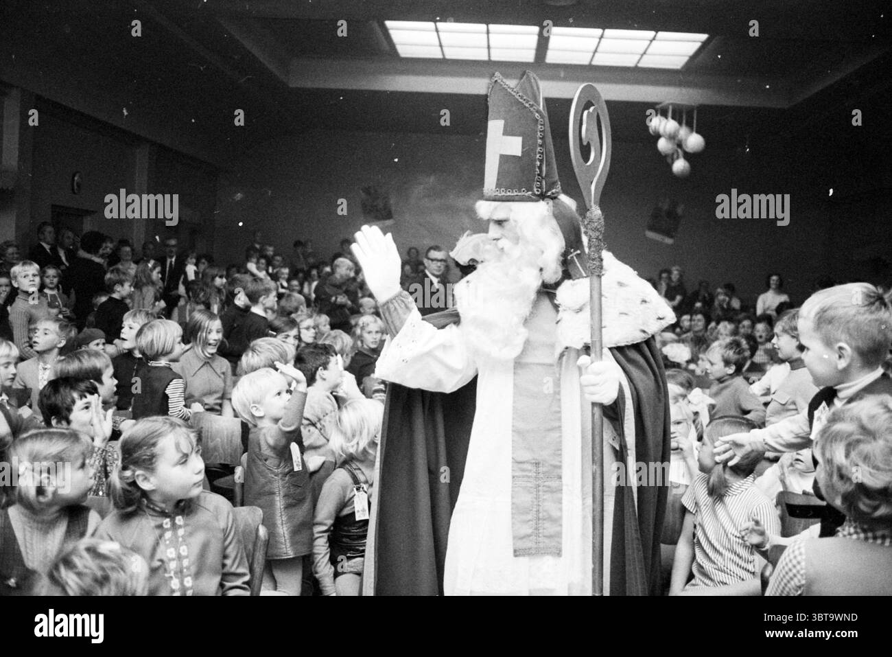 Sint in Leiden Noord Zuid Hollandse Vervoersmaatschappij N.V. NZ, Whizgle News, Dutch Desk, Paesi Bassi, 1950 - 2000 il 25-11-1970. L'immagine contiene questi argomenti. In un vivace ambiente interno, una folla di bambini riempie lo spazio, i loro volti sono un mix di eccitazione e curiosità. La scena irradia un'atmosfera festosa, probabilmente durante una festa. Al centro si trova una figura vestita con un grande abito cerimoniale, completo di un mantello scorrevole e di un alto cappello ornato con una croce. Questo personaggio ha un grande personale contorto, che contribuisce a creare un'aria di autorità e mistero. TH Foto Stock