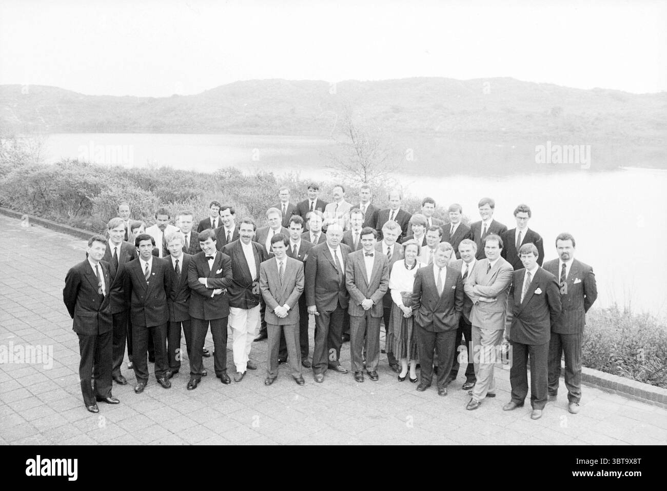 Gruppo alla degustazione di vini fa. Kwast Wijn, Whizgle News, Dutch Desk, Paesi Bassi, 1950 - 2000 il 09-05-1988. L'immagine contiene questi argomenti. La scena cattura un grande gruppo di individui disposti in modo un po' formale, in piedi in file ordinate. L'ambiente sembra essere un'area all'aperto vicino a un lago, con un corpo d'acqua calmo e riflettente che si estende sullo sfondo. Le dolci colline possono essere viste in lontananza, avvolte da una sottile macchia che aggiunge un senso di profondità alla scena. Il gruppo è principalmente vestito con abiti scuri, indicando un'occasione formale, con molti lacci. Il predo Foto Stock