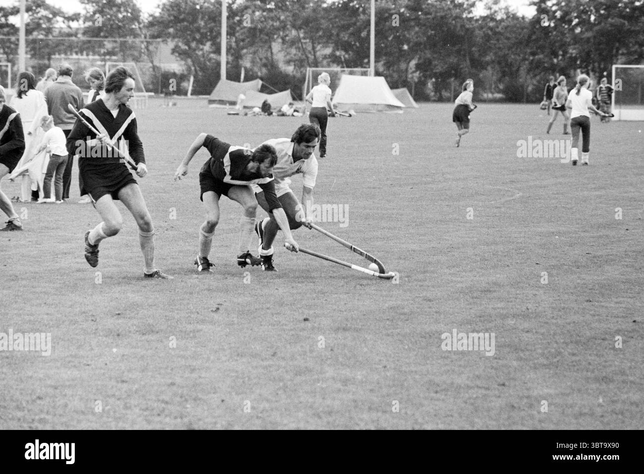 Torneo finale di hockey Alliance Hockey, Whizgle News, Dutch Desk, Paesi Bassi, 1950 - 2000 il 24-08-1980. Questi sono gli elementi dell'immagine. La scena cattura un momento dinamico su un grande campo erboso, dove si svolge un gioco vivace. In primo piano, tre individui sono impegnati in un gioco intenso, due di loro rivestiti in uniformi scure, mentre uno indossa un vestito di colore chiaro. Il giocatore al centro si sta piegando in basso, manovrando abilmente un bastone che si estende verso la palla, creando un senso di urgenza e azione. Intorno a loro, molti altri popolano il campo, partecipando Foto Stock