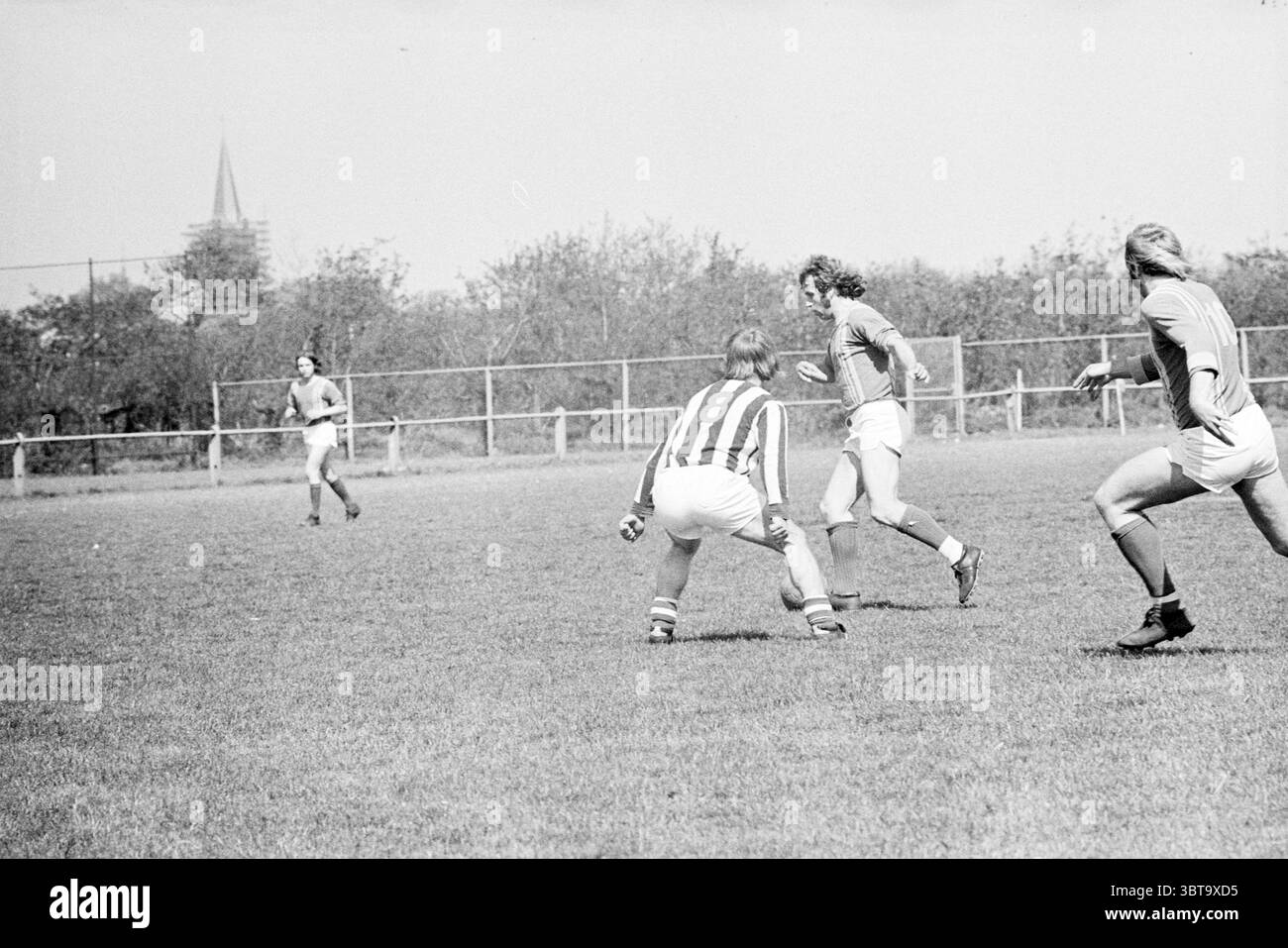 Una partita di calcio., Whizgle News, Dutch Desk, Paesi Bassi, 1950 - 2000. Questi sono gli elementi dell'immagine. La scena raffigura un momento vivace in una giornata di sole in un campo erboso, chiaramente predisposto per una partita di calcio. I giocatori sono sparsi in tutto il campo, ognuno con una divisa distinta, indicativa di due squadre in competizione. A sinistra, un giocatore con una maglia a strisce dribbla con abilità, concentrazione e dinamicità nella postura. La sua espressione significa determinazione mentre si dirige verso i suoi avversari. Alla sua destra, un compagno di squadra, rivestito in uniforme di tinta unita, è pronto a sostenerlo, con gli occhi ben fissati Foto Stock