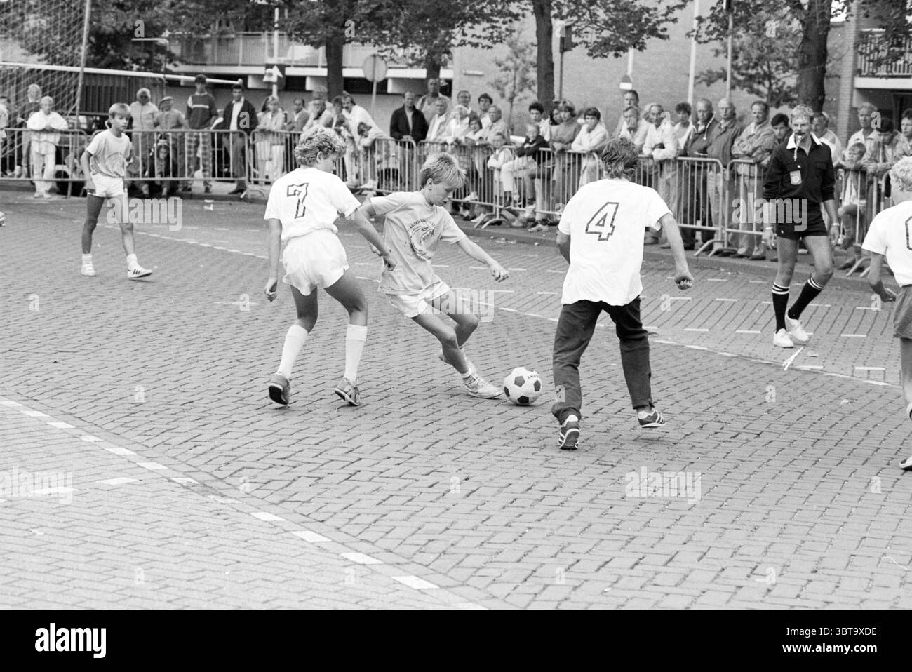Torneo di Street football Santpoort Street football Santpoort, Whizgle News, Dutch Desk, Paesi Bassi, 1950 - 2000 il 03-08-1986. Questi sono gli elementi dell'immagine. La scena cattura un momento dinamico di una partita di Street Soccer, piena di energia e movimento. Il terreno è contrassegnato da una superficie di mattoni a motivi geometrici, che fornisce uno sfondo testurizzato per l'azione. In primo piano, tre giovani giocatori sono impegnati in un gioco intenso. Un giocatore, vestito con una maglia di colore chiaro, sta manovrando con abilità la palla da calcio, mentre altri due, rivestiti con abiti bianchi e scuri a contrasto, si stanno chiudendo Foto Stock