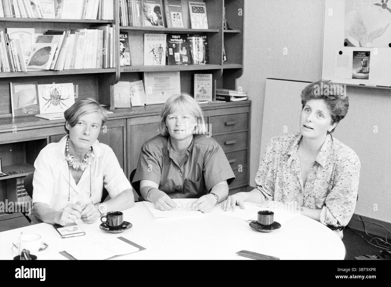Sig.ra Gottmer con colleghi Bloemendaal Printing House Printing Persons Bloemendaal, Whizgle News, Dutch Desk, Paesi Bassi, 1950 - 2000 il 07-07-1987. Questi argomenti sono mostrati nell'immagine. In una sala ben illuminata, tre donne sono sedute intorno a un tavolo circolare. Il tavolo è coperto da fogli di carta e alcune piccole tazze nere, suggerendo un incontro informale o una discussione. Lo sfondo è pieno di scaffali che espongono vari libri e riviste, creando un'atmosfera intellettuale. Le donne mostrano espressioni diverse, riflettendo una serie di emozioni, dalla contemplazione all'impegno. Uno Wo Foto Stock
