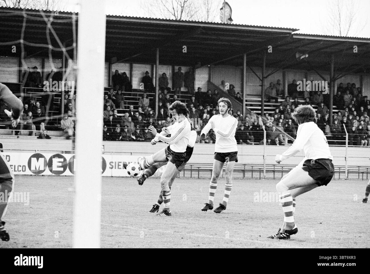 Haarlem - PEC Football Haarlem, Whizgle News, Dutch Desk, Paesi Bassi, 1950 - 2000 il 09-11-1975. L'immagine include questi argomenti. La scena cattura un momento intenso su un campo da calcio sullo sfondo di un modesto stadio sportivo. Due squadre, vestite con divise a contrasto, una con maglie bianche e pantaloncini scuri e l'altra con calze a righe sportive, sono impegnate in un gioco dinamico. In primo piano, un giocatore con una camicia bianca dimostra agilità mentre colpisce abilmente una palla da calcio, il suo corpo è inclinato per un contatto ottimale. La determinazione sul suo viso mette in evidenza la tensione del gioco. Nelle vicinanze, Foto Stock