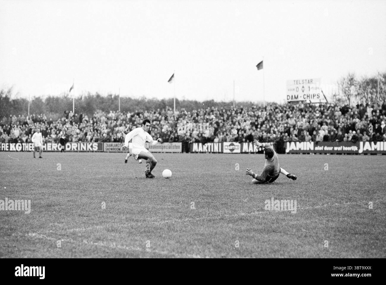 Football Telstar - NEC, Whizgle News, Dutch Desk, Paesi Bassi, 1950 - 2000 il 16-11-1969. Questi argomenti vengono visualizzati nell'immagine. La scena cattura un momento dinamico su un campo da calcio in erba. In primo piano, due giocatori si impegnano in un gioco intenso. Un giocatore, vestito con una divisa bianca, sta manovrando abilmente la palla, mentre l'altro, indossando l'uniforme di un portiere, è in immersione, braccia allungate, cercando di bloccare il colpo. Lo sfondo è pieno di una folla densamente affollata, i cui volti sono un mix di anticipazione ed eccitazione. Sono vestite con diverse giacche e cappelli, che sembrano essere brav Foto Stock
