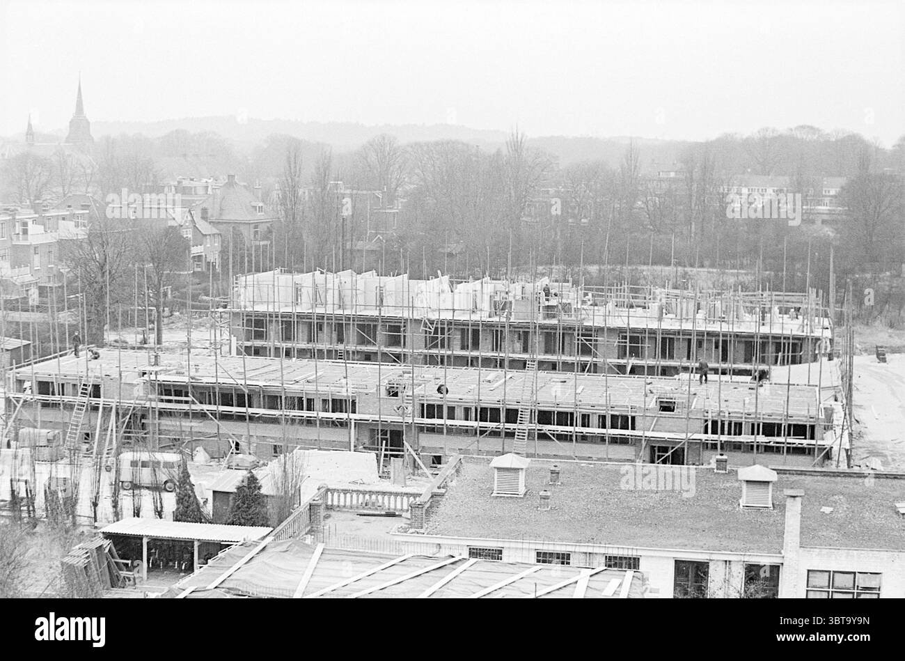 La nuova costruzione ospita Haarlem Nederland, Whizgle News, Dutch Desk, Paesi Bassi, 1950 - 2000 il 19-03-1964. Questi argomenti sono mostrati nell'immagine. La scena raffigura un cantiere pieno di attività. In primo piano, è visibile una struttura di una grande struttura, che si innalza in modo prominente con travi in acciaio e impalcature in legno creando uno schema simile a una griglia. Il profilo scheletrico suggerisce gli inizi di un edificio a più piani, con i lavoratori impegnati in vari compiti tra i supporti. Intorno alla struttura si trovano diversi tumuli di terra e pile di materiali, tra cui tavole e. Foto Stock