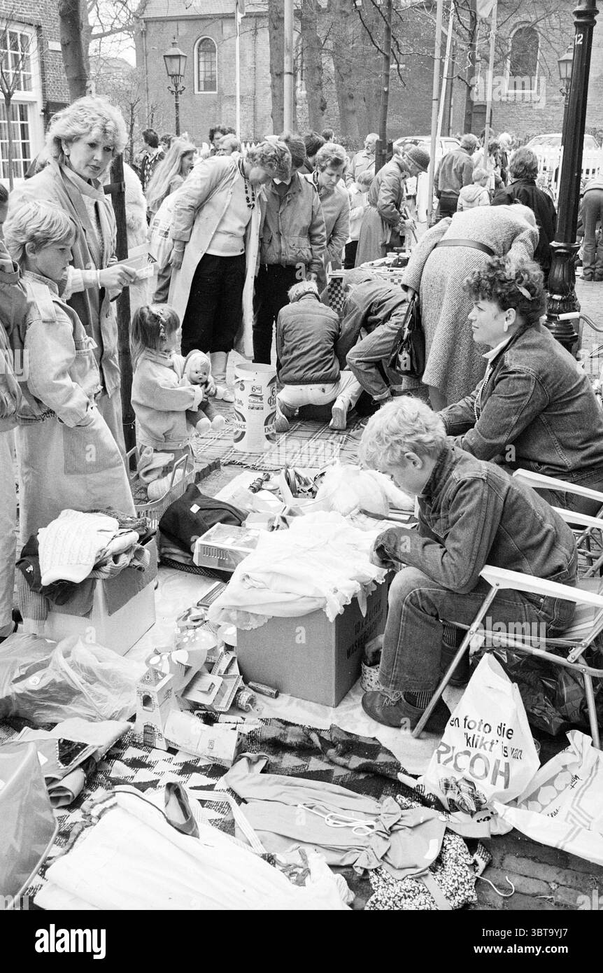 Braderie Oud Velsen, Whizgle News, Dutch Desk, Paesi Bassi, 1950 - 2000 il 05-05-1985. Questi argomenti sono mostrati nell'immagine. La scena e' movimentata di attivita', adagiata in una vivace area di mercato all'aperto. Numerosi individui sono impegnati in varie attività, creando un senso di comunità e interazione. In primo piano, una donna si siede su una sedia pieghevole, girando attraverso scatole piene di articoli assortiti come vestiti e giocattoli. Ha i capelli ricci e appare concentrata sui suoi reperti, occasionalmente guardando l'ambiente circostante. Nelle vicinanze, un bambino piccolo, forse con i capelli biondi, è affascinato da Foto Stock