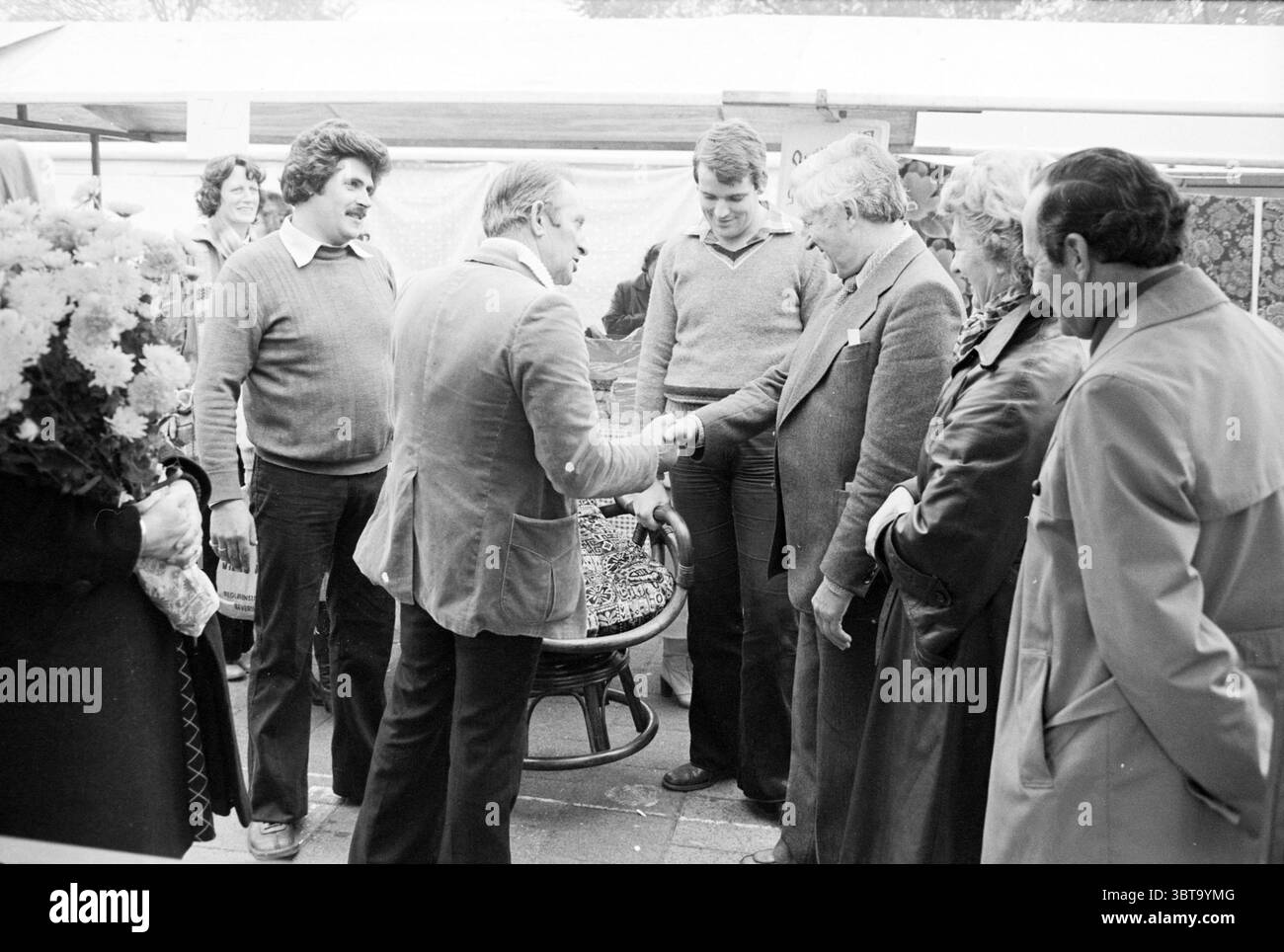 Il mercante di mercato dice addio B'wijk Markt, Whizgle News, Dutch Desk, Paesi Bassi, 1950 - 2000 il 01-11-1978. Questi sono gli argomenti nell'immagine. In una vivace scena di mercato piena di energia vivace, diverse figure sono impegnate in una conversazione intorno a un tavolo circolare che mostra una serie di merci. L'attenzione centrale è rivolta a due uomini, uno con una giacca di colore chiaro e l'altro con un maglione, che stringono la mano, simboleggiando un accordo o un saluto amichevole. Intorno a loro ci sono altri tre individui, ognuno che trasuda una personalità diversa. A sinistra, una donna in abiti scuri regge un bouquet di Foto Stock