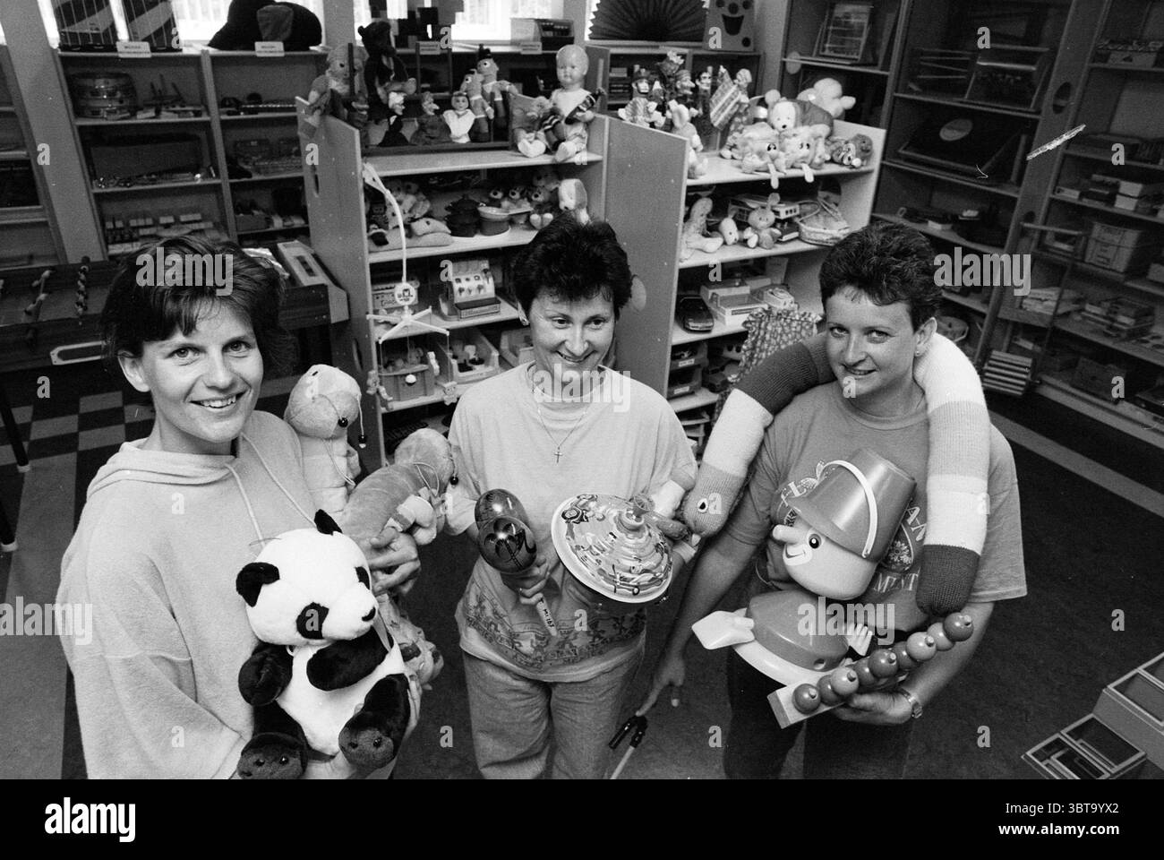 Speel-o-Theek Hartekamp, Whizgle News, Dutch Desk, Paesi Bassi, 1950 - 2000 il 20-06-1990. L'immagine mostra questi argomenti. In un ambiente vivace e vivace pieno di vari giocattoli e oggetti per bambini, tre persone stanno in primo piano, ognuno con un assortimento di oggetti colorati. Lo sfondo è una sala giochi ben fornita o un negozio di giocattoli, con scaffali rivestiti di peluche, bambole e giochi, che creano un'atmosfera vivace. I tre individui, che appaiono impegnati e felici, presentano una calda atmosfera comunitaria. Sono vestiti in modo informale, con texture morbide e colori chiari e neutri Foto Stock