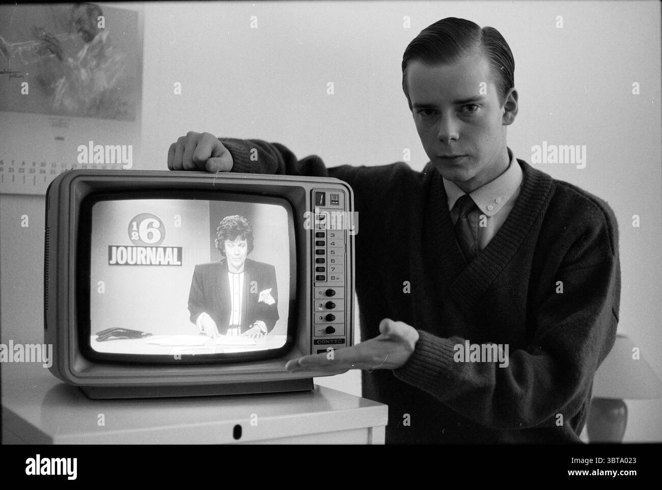 Erwin Segaar su TV People Television, Whizgle News, Dutch Desk, Paesi Bassi, 1950 - 2000 il 06-05-1988. L'immagine include questi argomenti. In una scena monocromatica, un giovane si trova con fiducia accanto a un televisore d'epoca. Ha i capelli ben curati e indossa un maglione scuro su una camicia con colletto, incarnando un'estetica retrò. La sua espressione è seria ma coinvolgente mentre si dirige verso lo schermo televisivo, dove è seduto un presentatore di notizie, riportando animatamente. Il presentatore, anche catturato nei toni silenziosi, indossa un abito e ha un comportamento mirato, aggiungendo un livello di autorità al Foto Stock