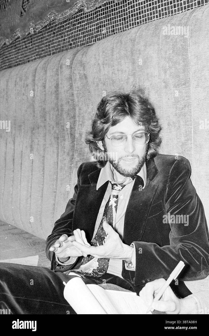 Steven Bishop cantante chitarrista Music, Whizgle News, Dutch Desk, Paesi Bassi, 1950 - 2000 il 16-03-1977. Questi argomenti sono mostrati nell'immagine. Un uomo con capelli leggermente ondulati e di media lunghezza siede su un divano imbottito. Ha una barba ben curata e indossa occhiali con una montatura sottile. Il suo outfit è composto da un blazer scuro e morbido che riflette una sottile lucentezza, abbinato a una cravatta a motivi geometrici che aggiunge un tocco di vivacità. Il tessuto del divano appare ricco e testurizzato e sfoggia un tono tenue che si abbina all'abbigliamento maschile. Dietro di lui, un design astratto è sottilmente incorporato nello sfondo, suggerisci Foto Stock