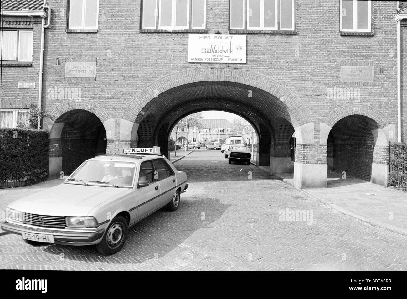 Quartiere in vista: Quartieri storici Haarlem Kloppersingel Paesi Bassi, Whizgle News, Dutch Desk, Paesi Bassi, 1950 - 2000 il 27-04-1981. L'immagine contiene questi argomenti. La scena presenta un arco in mattoni, caratterizzato dalle sue robuste texture rosso-marrone, che conduce in una strada fiancheggiata da edifici residenziali. L'arco stesso è ampio e invitante, con due archi distinti che consentono il passaggio sotto di esso. I mattoni sono intemprati, indicando l'età e aggiungendo al carattere della struttura. In primo piano, un'auto di colore chiaro, probabilmente in una sfumatura di grigio o beige, lo è Foto Stock