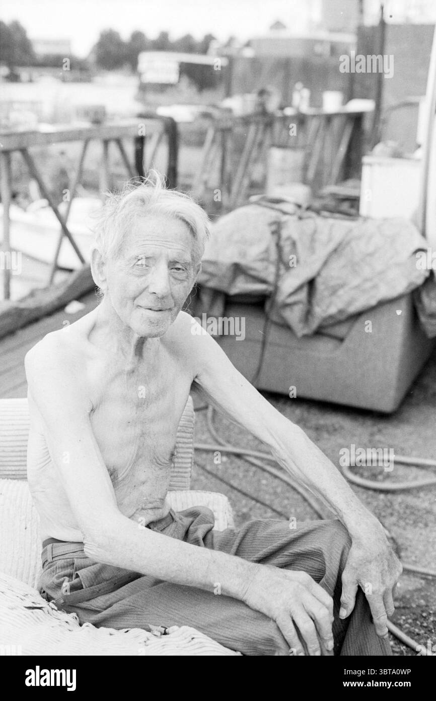 Man in Caravan SP., Whizgle News, Dutch Desk, Paesi Bassi, 1950 - 2000 il 08-08-1994. L'immagine include questi argomenti. In questa scena, un uomo anziano siede con un'espressione calma sul viso, trasudando un senso di resilienza e saggezza. La sua pelle è intemprata, suggerendo una vita piena di esperienze, e la sua sottile cornice suggerisce in età avanzata. Ha un sorriso gentile, che aggiunge un tocco di calore al suo comportamento. Lo sfondo è un po' sfocato ma rivela un ambiente rustico, caratteristico di un ambiente di lavoro con accenni di elementi industriali. Sono visibili vari oggetti, come strumenti e teloni, che creano Foto Stock