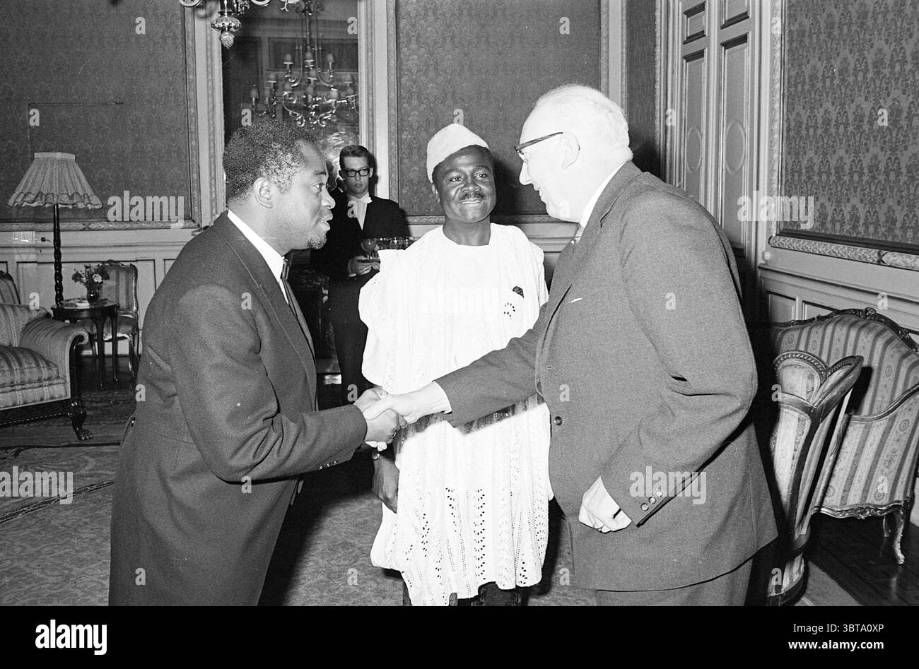 Reception Repubblica del Camerun. Atangana Hotel de Indes Recetions, Whizgle News, Dutch Desk, Paesi Bassi, 1950 - 2000 il 01-10-1964. Questi sono gli argomenti nell'immagine. Nella scena, due uomini sono impegnati in una stretta di mano ferma, un gesto che trasmette cameratismo e rispetto. Un uomo, vestito con un abito scuro, ha un'espressione attenta mentre si piega leggermente in avanti, indicando il coinvolgimento nella conversazione. L'altro uomo, che indossa un tradizionale ensemble bianco con dettagli intricati, si trova tra di loro, trasudando una presenza tranquilla. La camera stessa è arredata in modo opulento e presenta pareti intricate Foto Stock