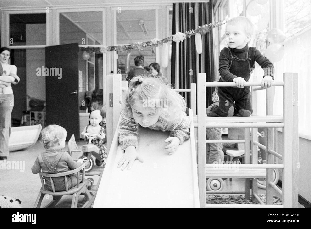 Open House del playgroup Het Piraatje Open Day e open House, Whizgle News, Dutch Desk, Paesi Bassi, 1950 - 2000 il 12-11-1977. Questi argomenti sono mostrati nell'immagine. La scena è piena della vivace energia dei bambini impegnati nel gioco all'interno di una spaziosa area interna. Una luce soffusa e diffusa inonda la stanza attraverso le grandi finestre, creando un'atmosfera calda e invitante. Le pareti sono adornate con decorazioni colorate che esaltano l'atmosfera gioiosa. In primo piano, due bambini sono il punto focale. Un bambino, una ragazza con i capelli ricci, è posizionato su un piccolo scivolo, la sua espressione è determinata Foto Stock