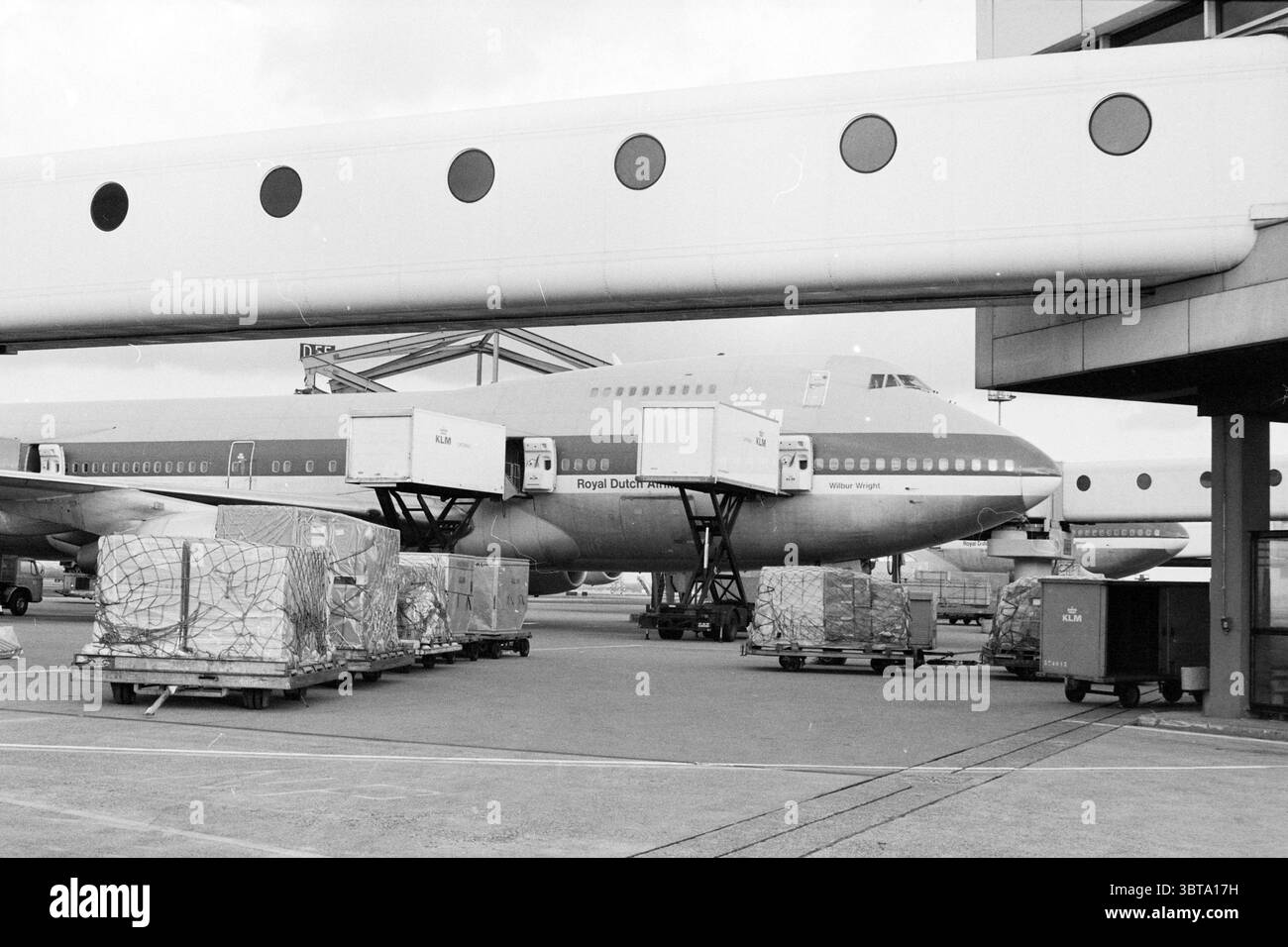Aerei e cargo Schiphol aereo Louis Bleriot, Whizgle News, Dutch Desk, Paesi Bassi, 1950 - 2000 il 21-12-1984. Questi sono gli argomenti nell'immagine. La scena cattura un vivace ambiente aeroportuale, caratterizzato da un grande aereo, probabilmente un Boeing 747, di stanza in un terminal. L'aereo presenta un design classico, prominente con il suo naso arrotondato e diversi motori, che mostra la sua livrea blu reale e bianca con accenti rossi. In primo piano, diversi container di carico sono impilati ordinatamente su pallet di grandi dimensioni, a indicare le attività incentrate sulle merci e sulla logistica. Questi container Foto Stock