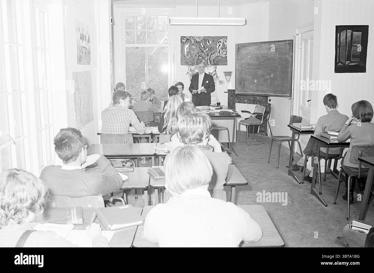 Rep. IVO School Bloemendaal Schools Bloemendaal Korte Parkweg, Whizgle News, Dutch Desk, Paesi Bassi, 1950 - 2000 il 12-01-1970. L'immagine contiene questi argomenti. La scena presenta un'aula piena di studenti seduti a banchi di legno individuali disposti in file, rivolti verso un'area anteriore dove si trova un insegnante. L'insegnante, vestito con un abito, è impegnato con gli studenti, che sembrano ascoltare intensamente. Le loro posture variano da inclinarsi leggermente in avanti a poggiare la testa sulle mani, indicando un mix di interesse e concentrazione. La camera è caratterizzata da alti soffitti Foto Stock