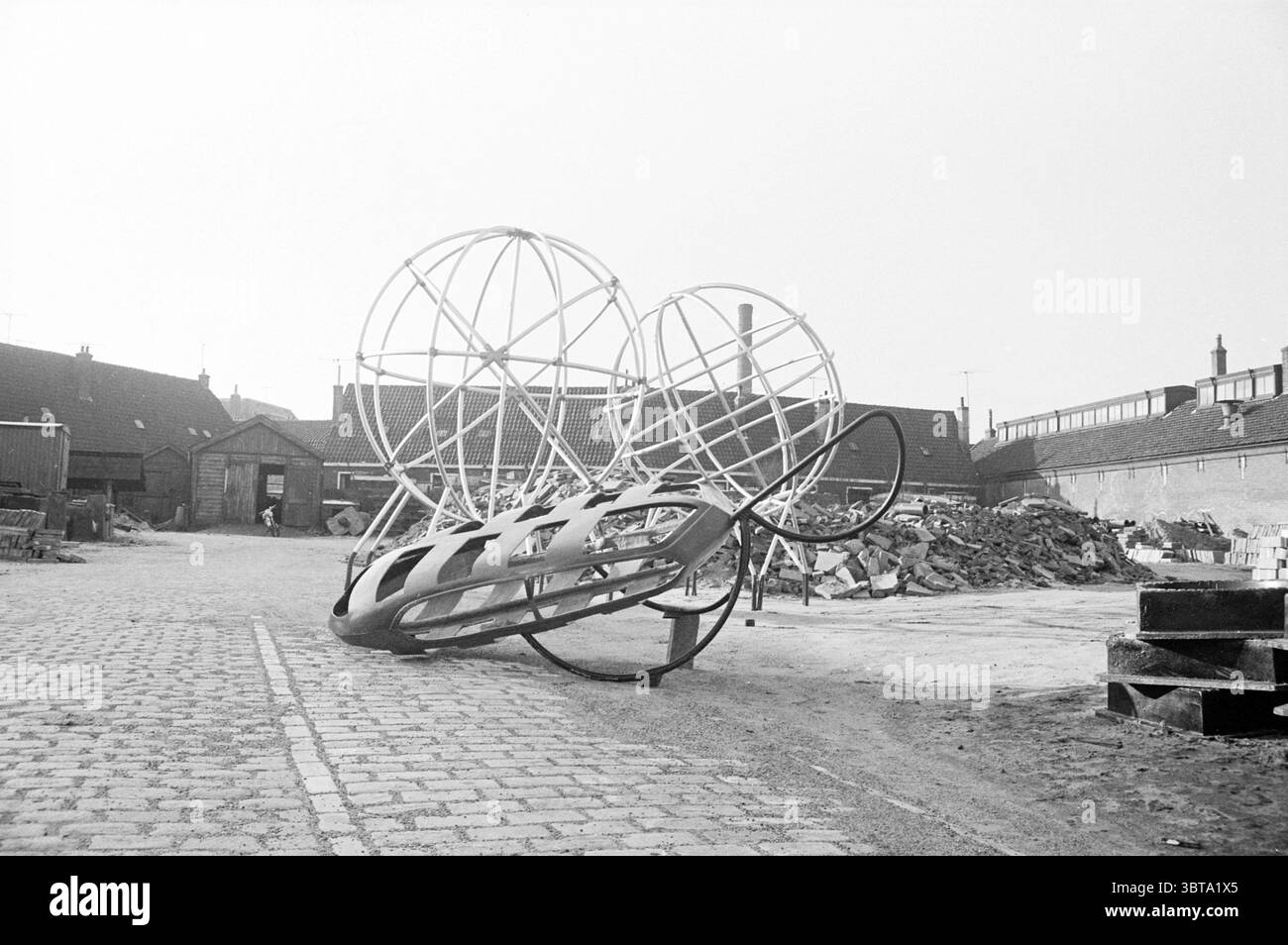Attrezzatura da arrampicata sul deposito, Whizgle News, Dutch Desk, Paesi Bassi, 1950 - 2000 su 12-1963. Questi sono gli elementi dell'immagine. Nella scena, una grande scultura astratta domina il primo piano, caratterizzata da una cornice metallica che assomiglia a una struttura stravagante e sovradimensionata. Il suo design è costituito da linee sinuose ed elementi circolari che creano un senso di movimento e giocosità. La base della scultura poggia su una grande ruota, contribuendo alla sua posizione dinamica. L'ambiente è un'area aperta, un po' industriale, con ciottoli che formano un sentiero che conduce verso un collettio Foto Stock