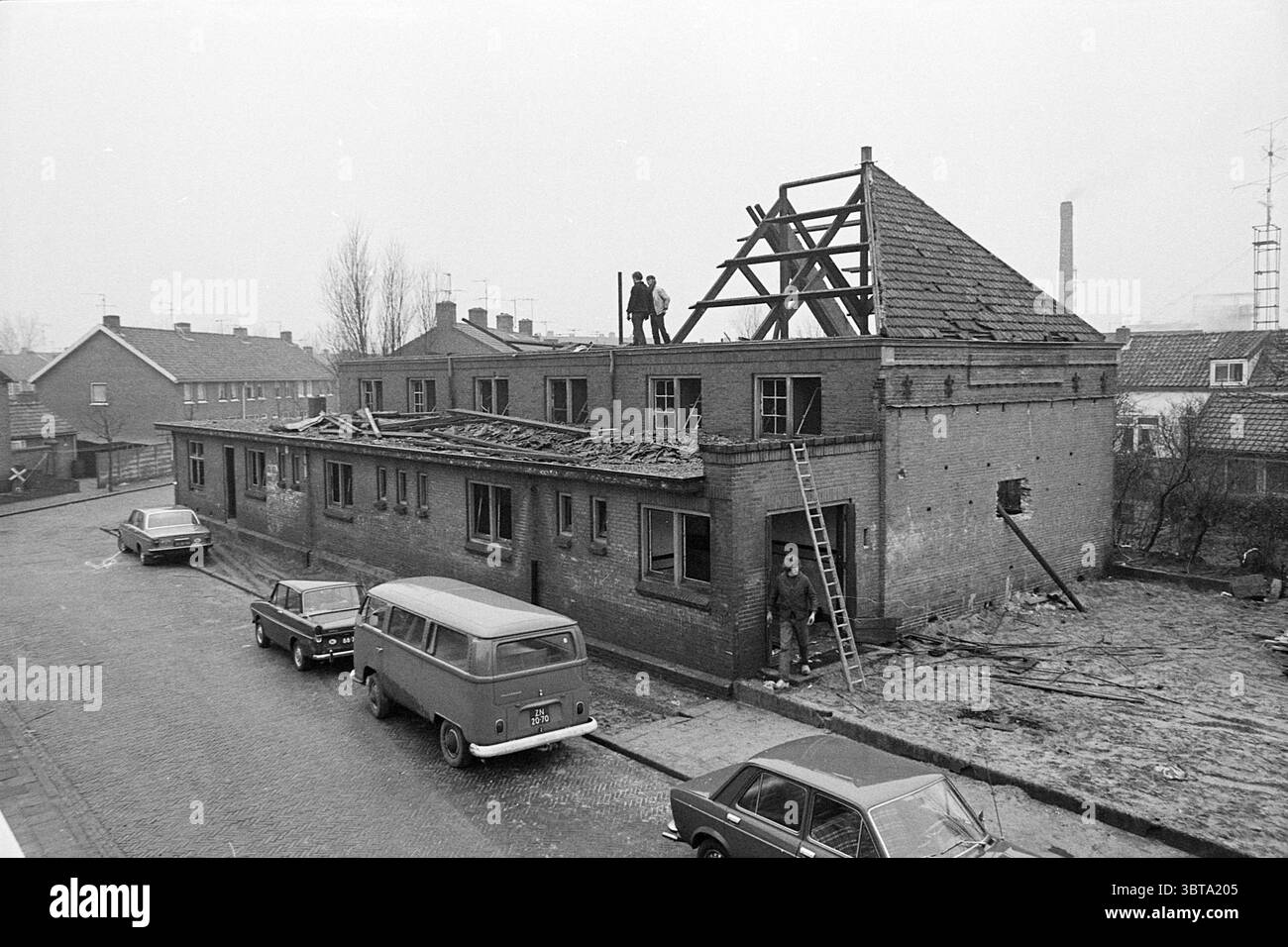 Edificio demolizione, Whizgle News, Dutch Desk, Paesi Bassi, 1950 - 2000 il 01-1972. Questi argomenti vengono visualizzati nell'immagine. La scena raffigura un cantiere con un edificio parzialmente demolito, che mostra la sua struttura del tetto incompiuta. L'edificio stesso e' fatto di mattoni, con alcune sezioni del tetto esposte, rivelando travi e supporti in legno. Due persone sono visibili in cima alla struttura, apparentemente impegnate nel lavoro, mentre un altro individuo si trova alla base della scala, osservando o assistendo. In primo piano, una strada stretta è costeggiata da diversi veicoli parcheggiati, tra cui un classico V Foto Stock